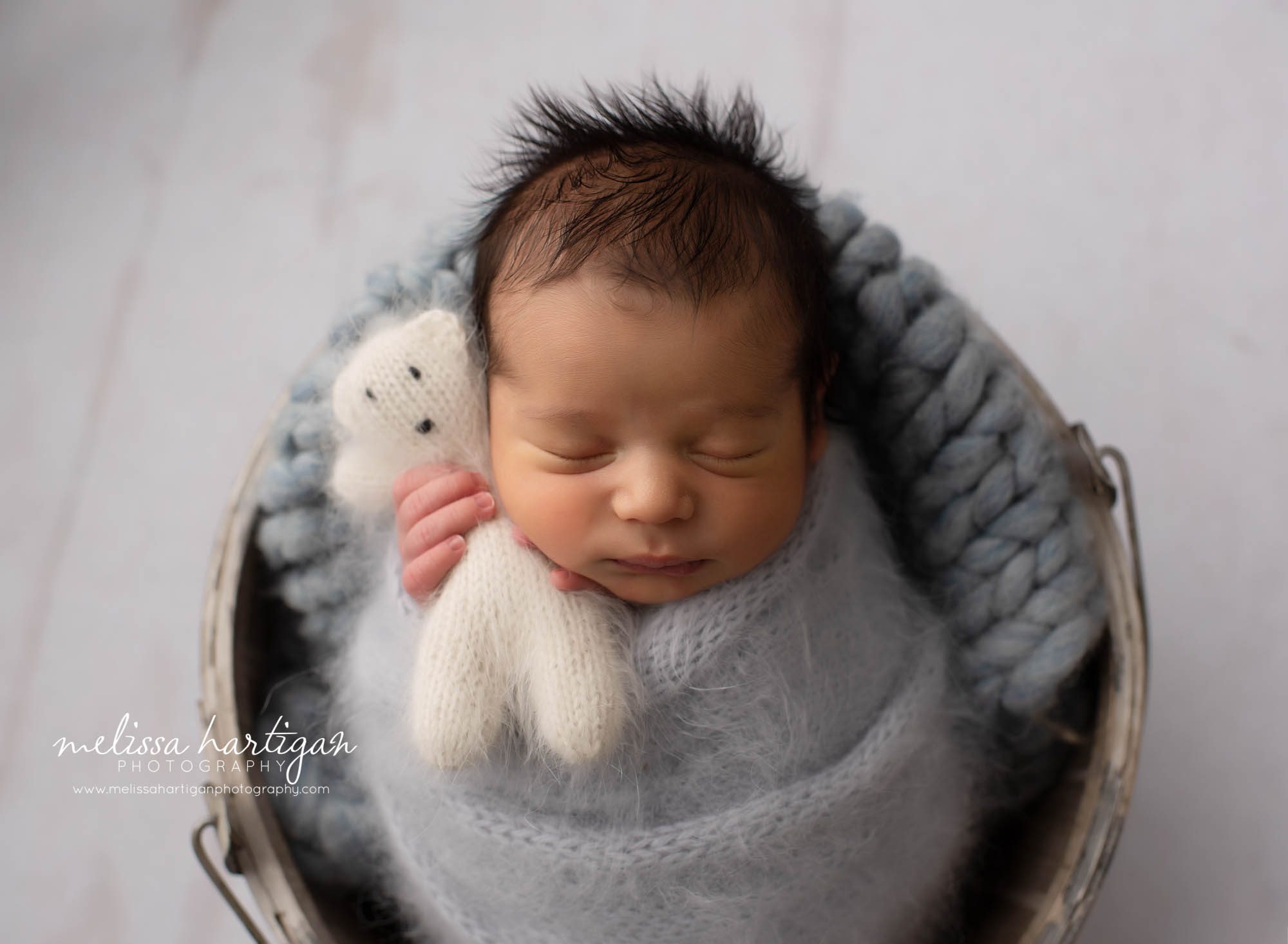 newborn boy wrapped in knitted gray wrap with cream colored knitted teddy bear posed in bucket