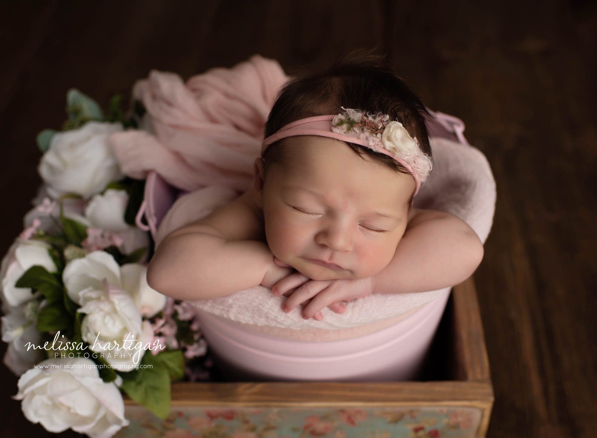 newborn baby girl posed with chin on hands in pink metal bucket inside wooden box with pink wrap draped over baby and white flower stems hartford County Newborn Photographer
