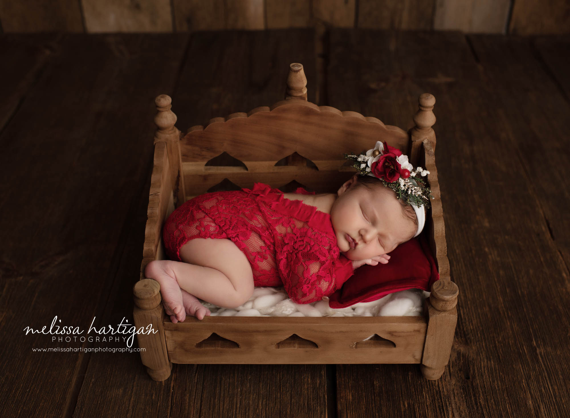 baby girl posed in wooden bed prop with red lace newborn romper and red green and white headband in holiday set up