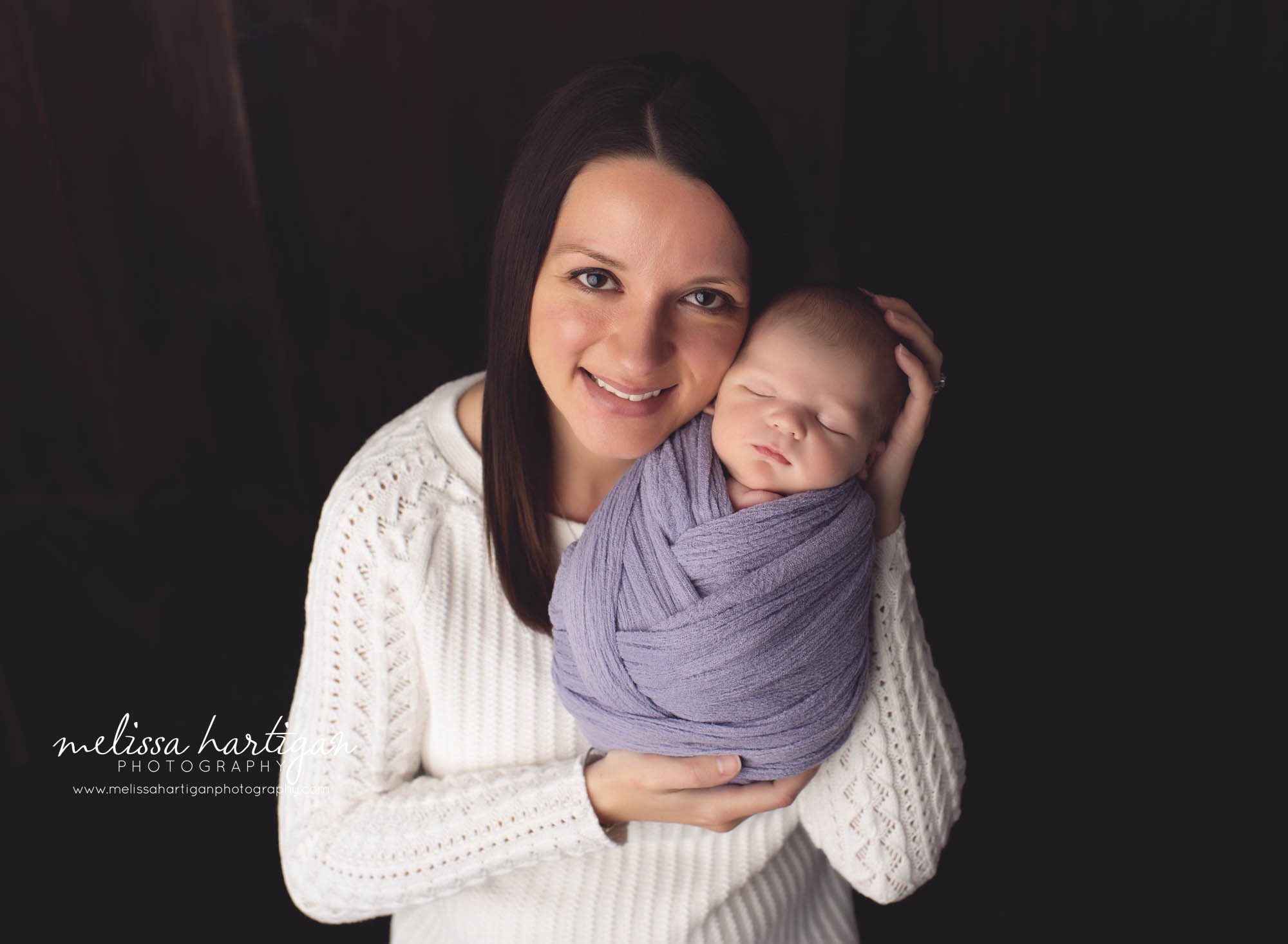 mom holding newborn girl wrapped in purple wrap smiling looking at camera
