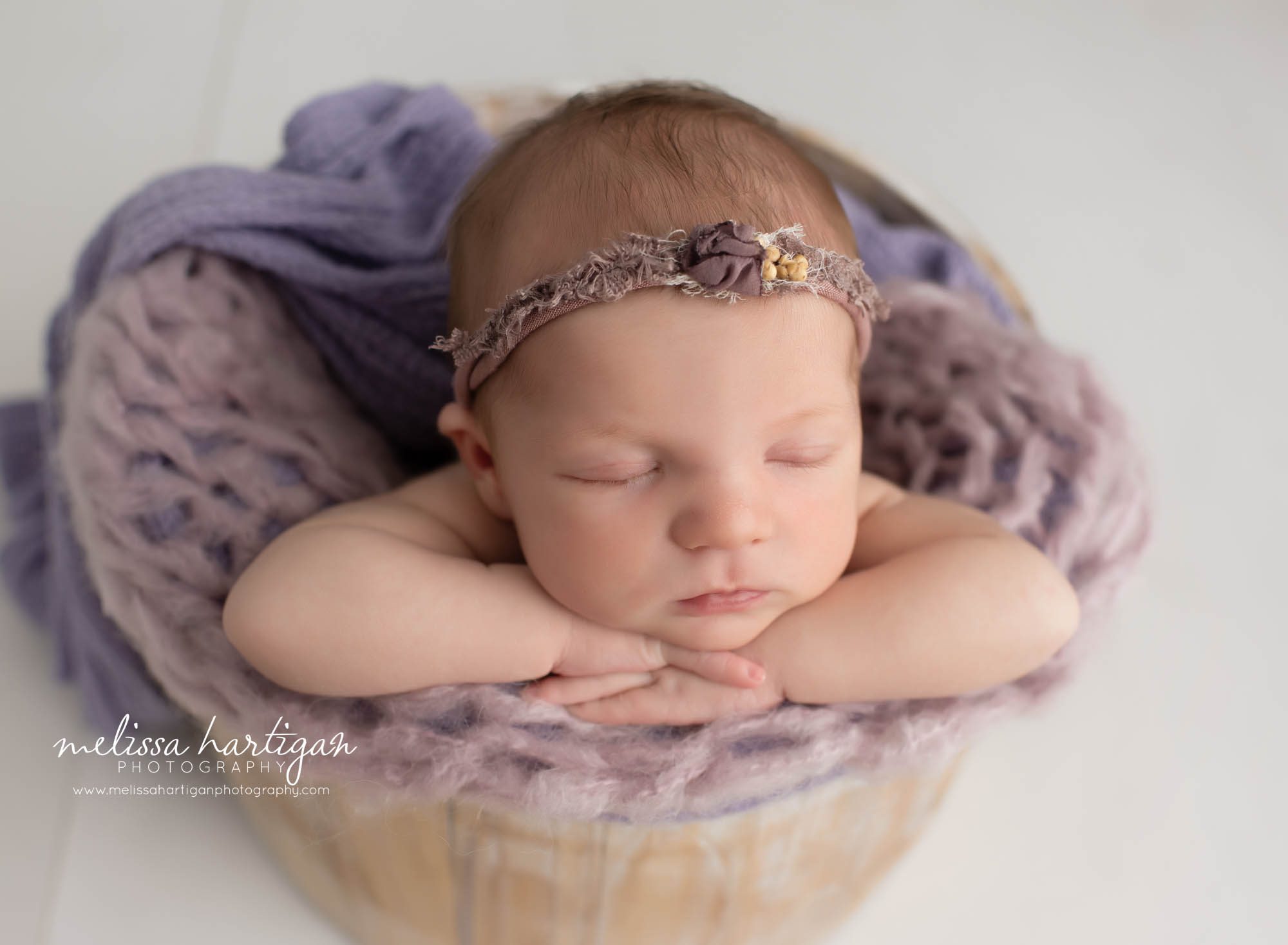 newborn baby girl posed in wooden bucket with purple chunky knitted layer and purple wrap chin resting on hands pose newborn photography