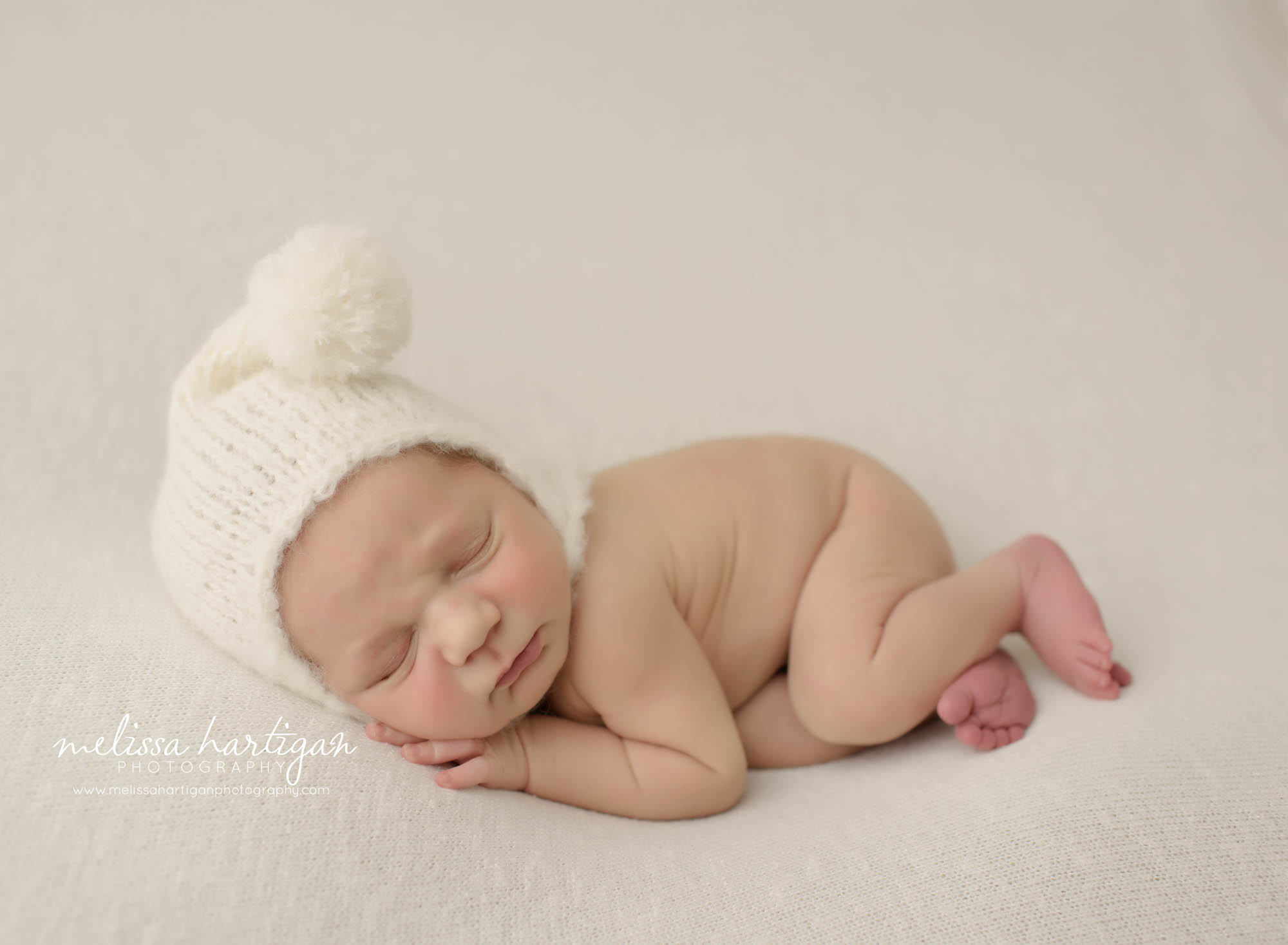 newborn baby sleeping on side with cream colored pom pom sleepy hat