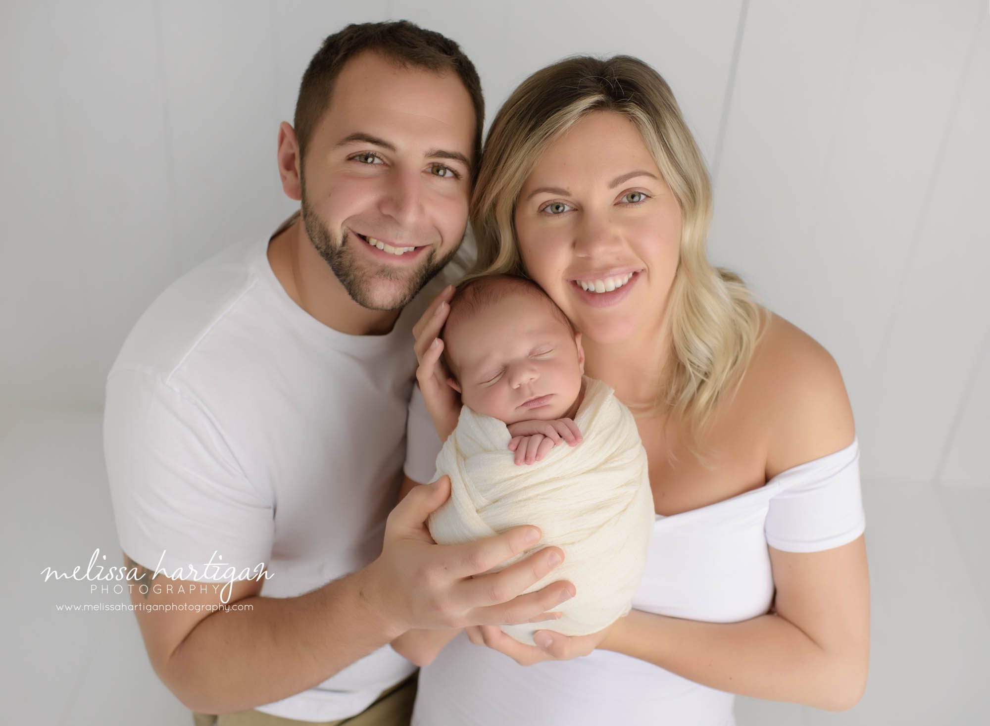 mom dad holding newborn son wrapped in ivory wrap in family photo in baby photography session in Connecticut studio