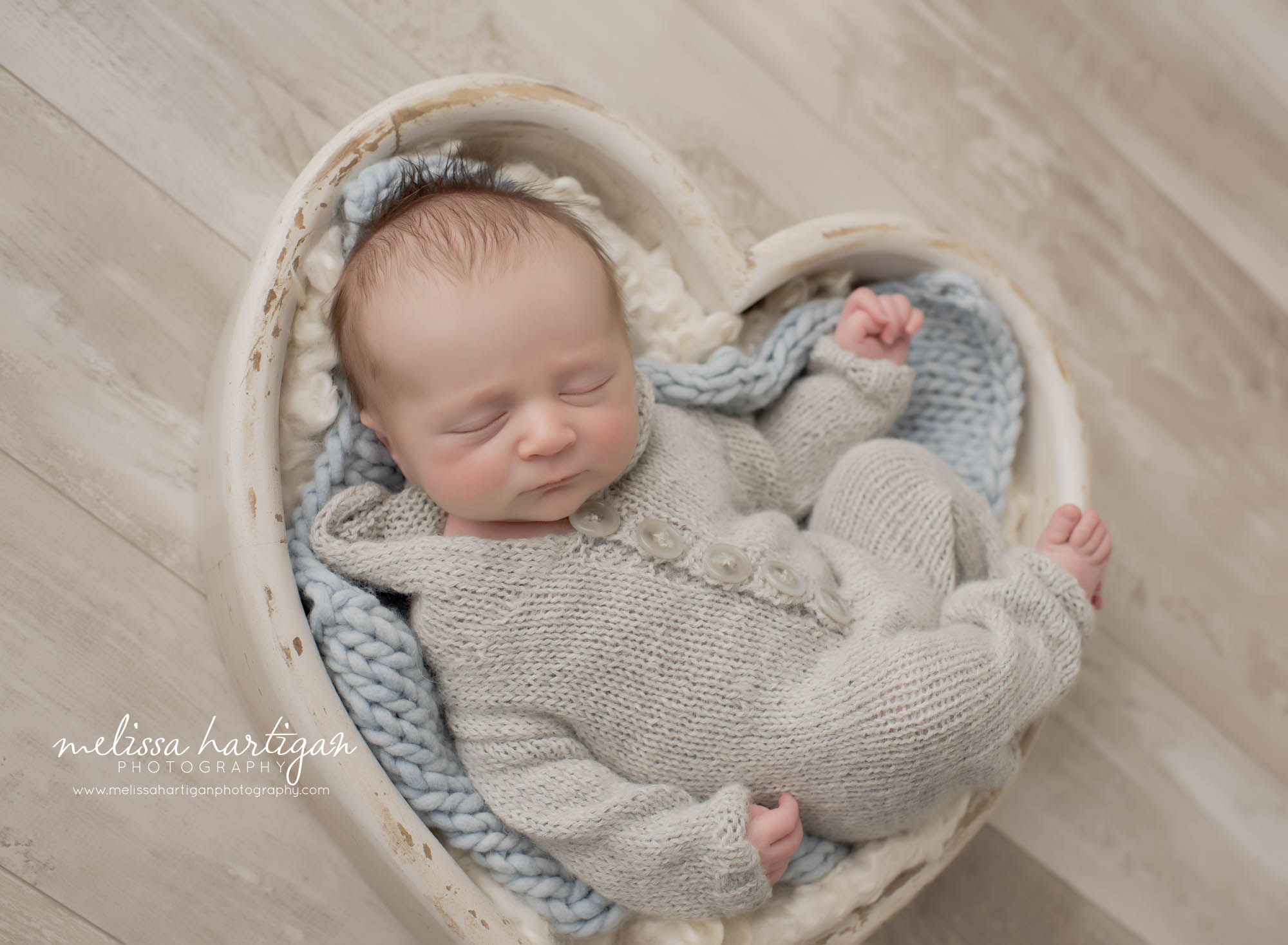 newborn boy posed in cream wooden heart prop light blue knit layer wearing light gray knitted baby outfit romper with hood