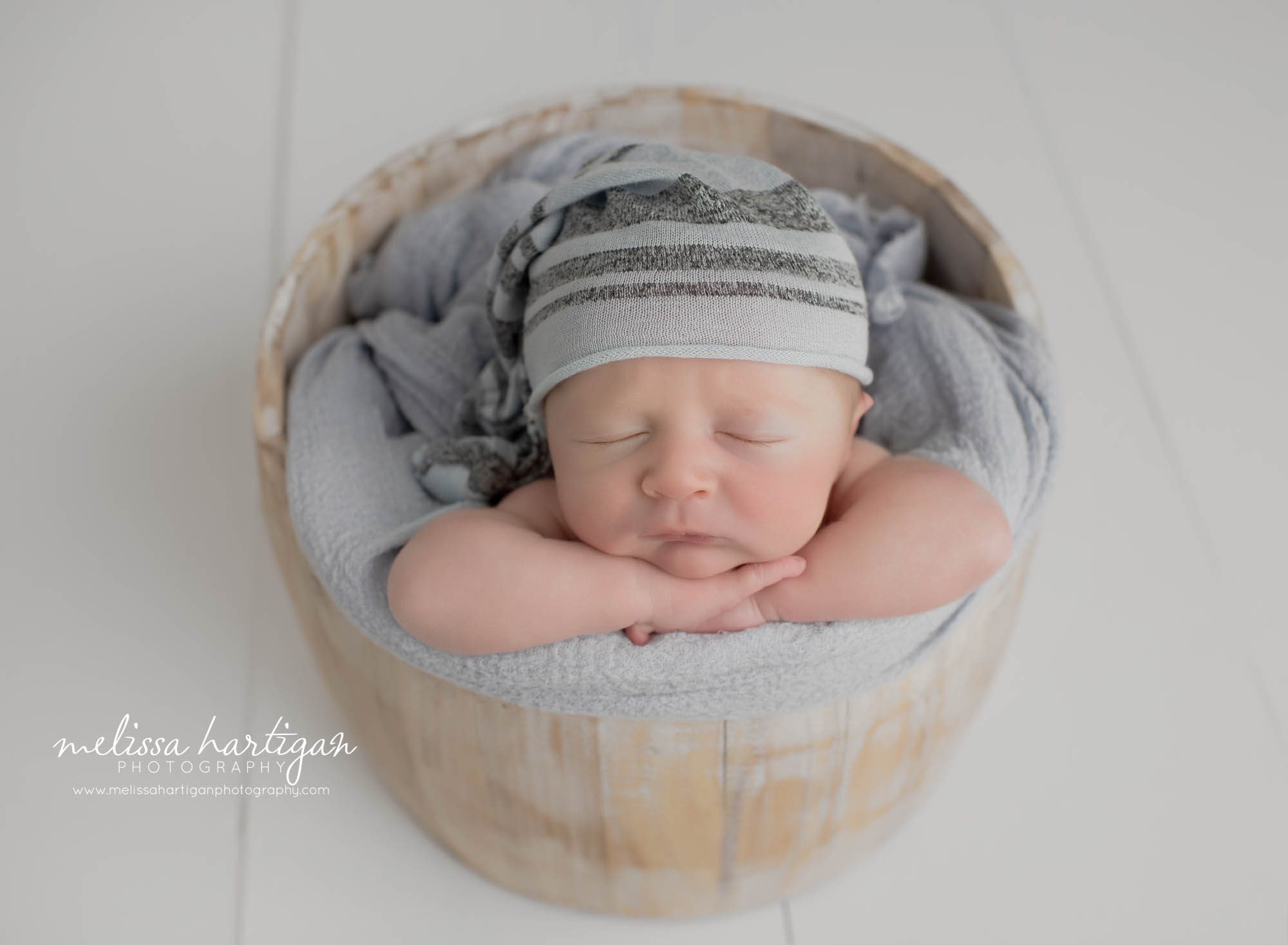 newborn boy posed in wooden bucket with light gray sleepy cap with stripes Tolland Newborn Photographer