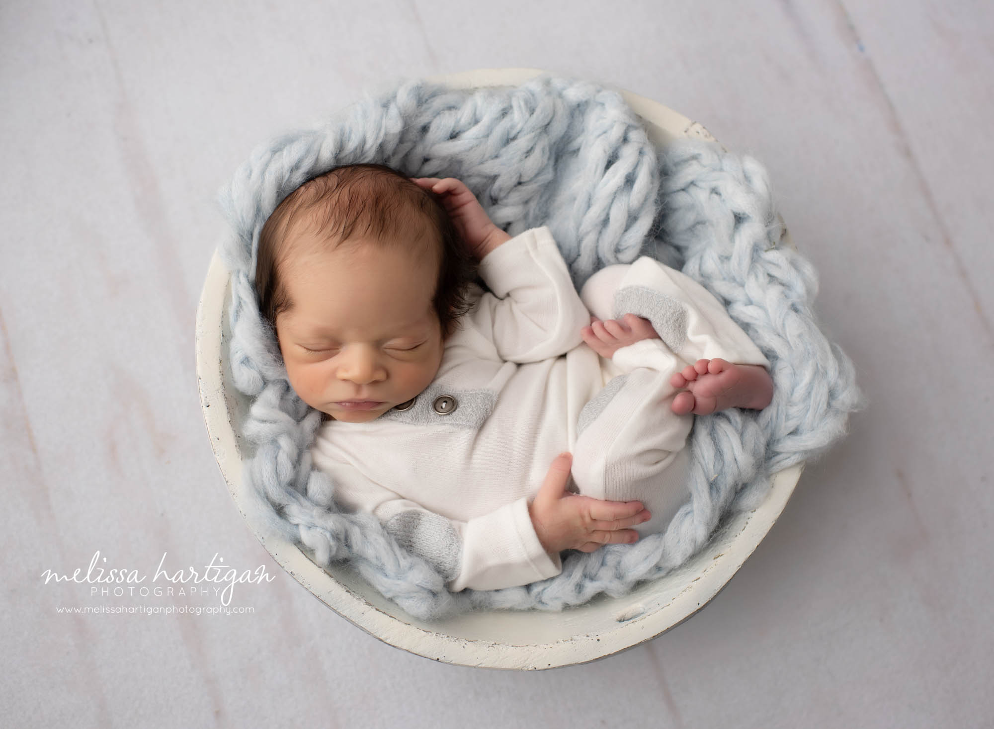 newborn baby boy posed in cream wooden bowl with light blue knitted layer wearing cream colored newborn outfit