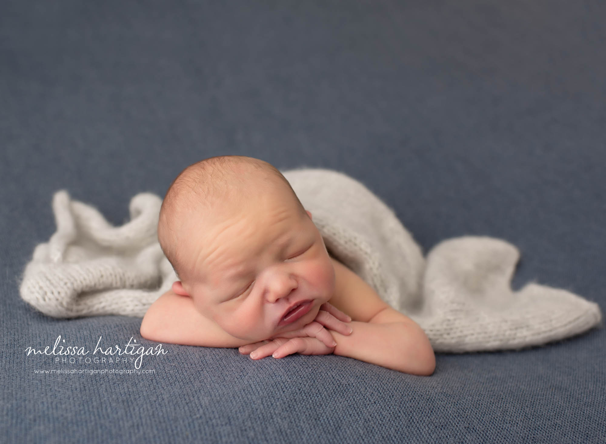 Newborn baby boy posed with chin on hands facing forward on blue gray backdrop with light gray knitted wrap drapped over