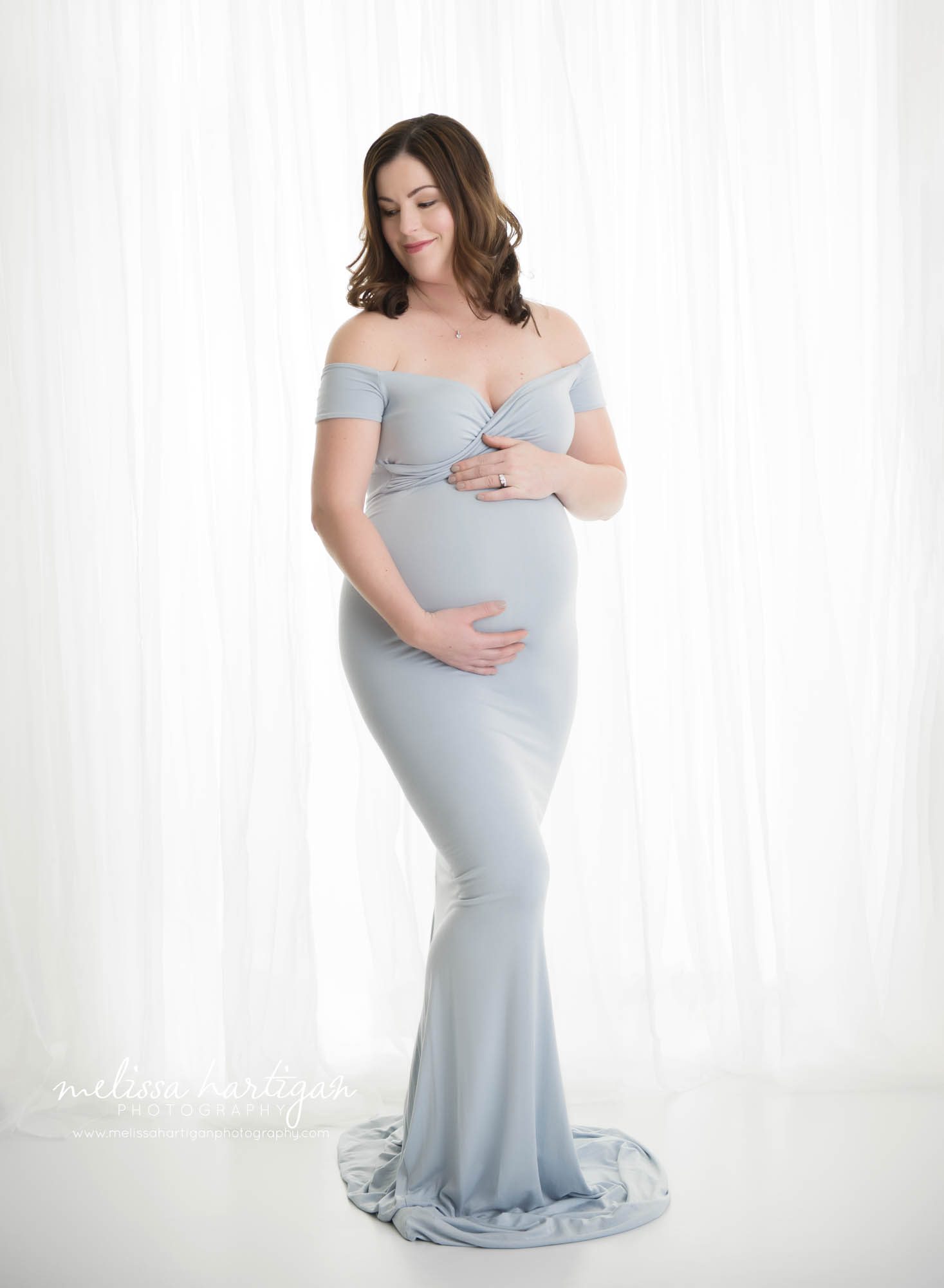 expectant mom standing holding baby bump wearing long form fitted blue dress against white backdrop