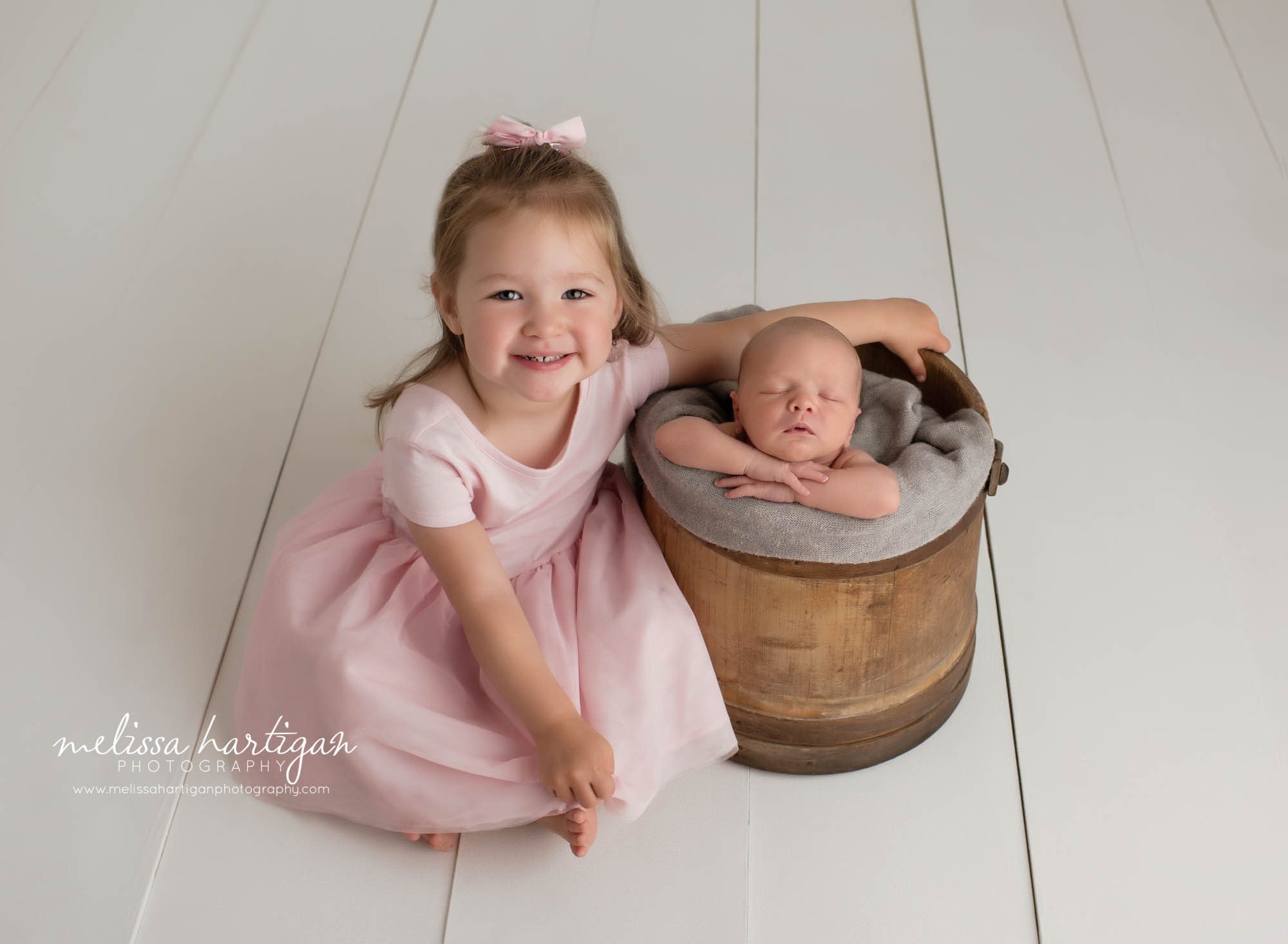 newborn baby boy posed in wooden bucket with big sister in pink dress sitting beside bucket