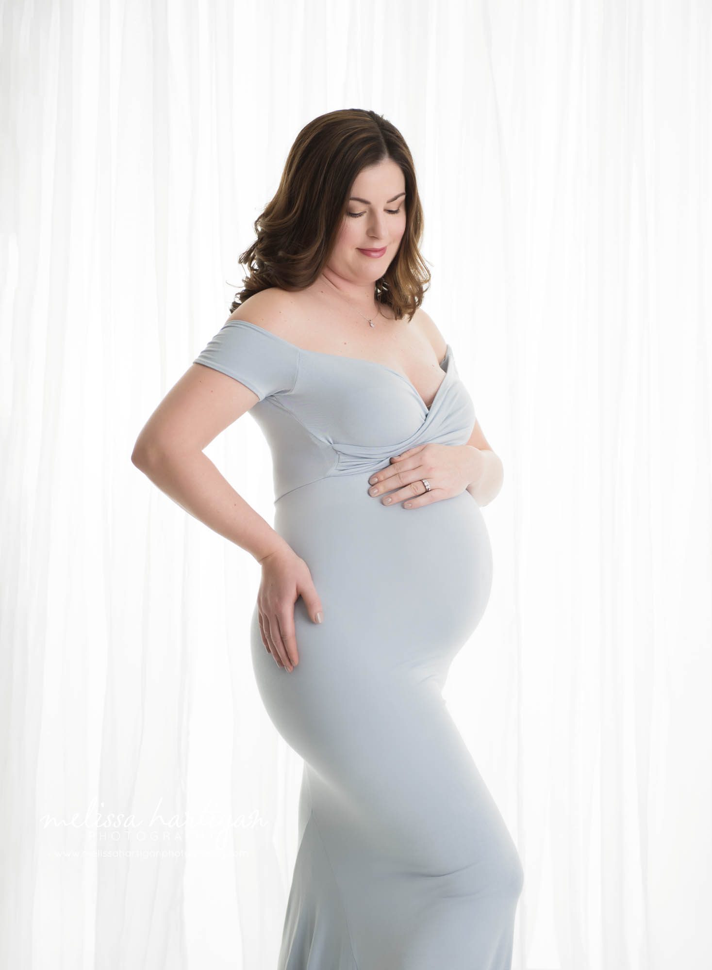 Expectant mom standing against white sheer curtain backdrop wearing off the shoulder long form fitting blue dress sweetheart neckline one hand on side one hand on baby bump looking down at baby bump Tolland Maternity Photography