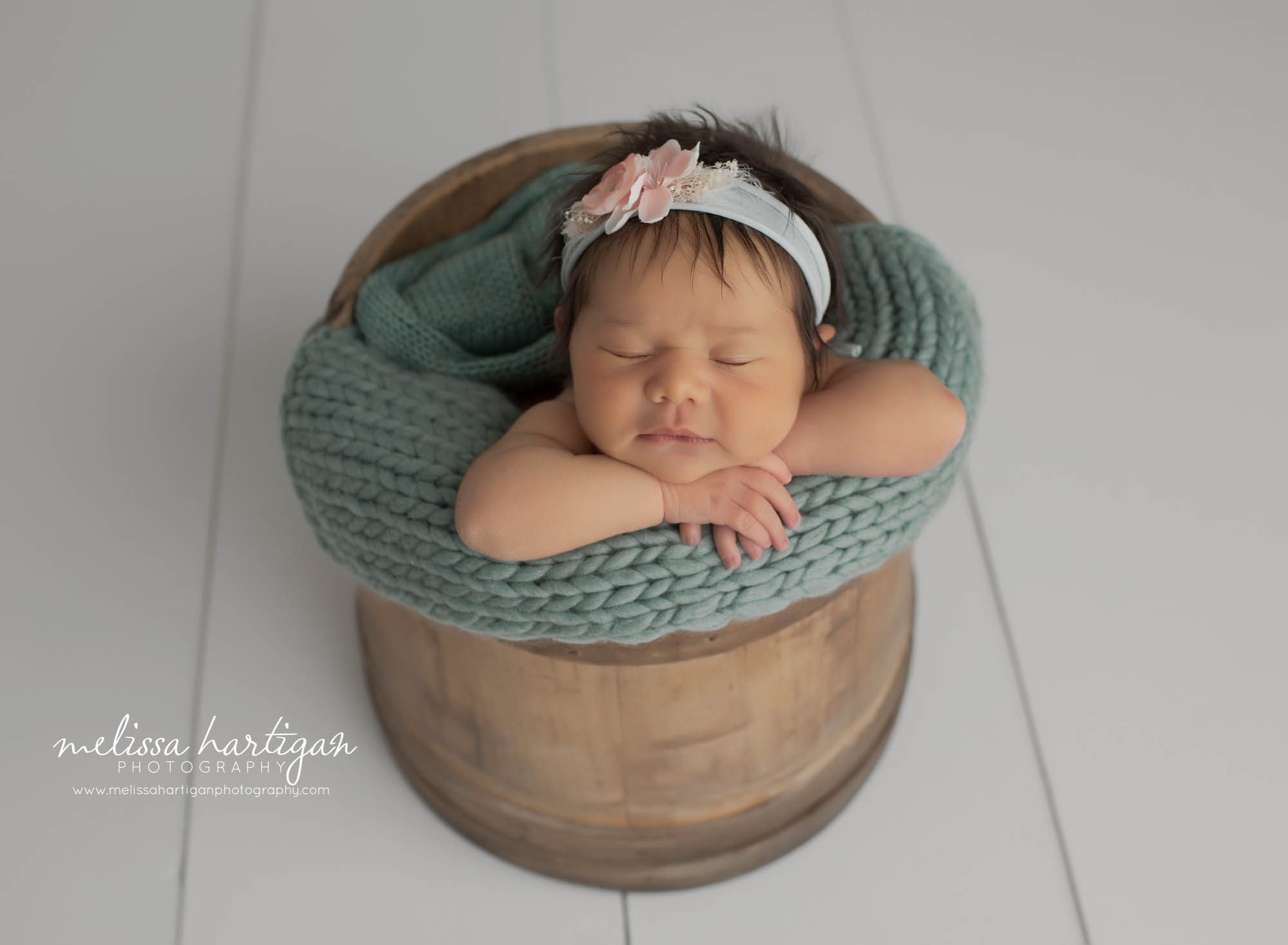 newborn baby girl posed in wooden bucket with dark sea green knitted layer and flower headband