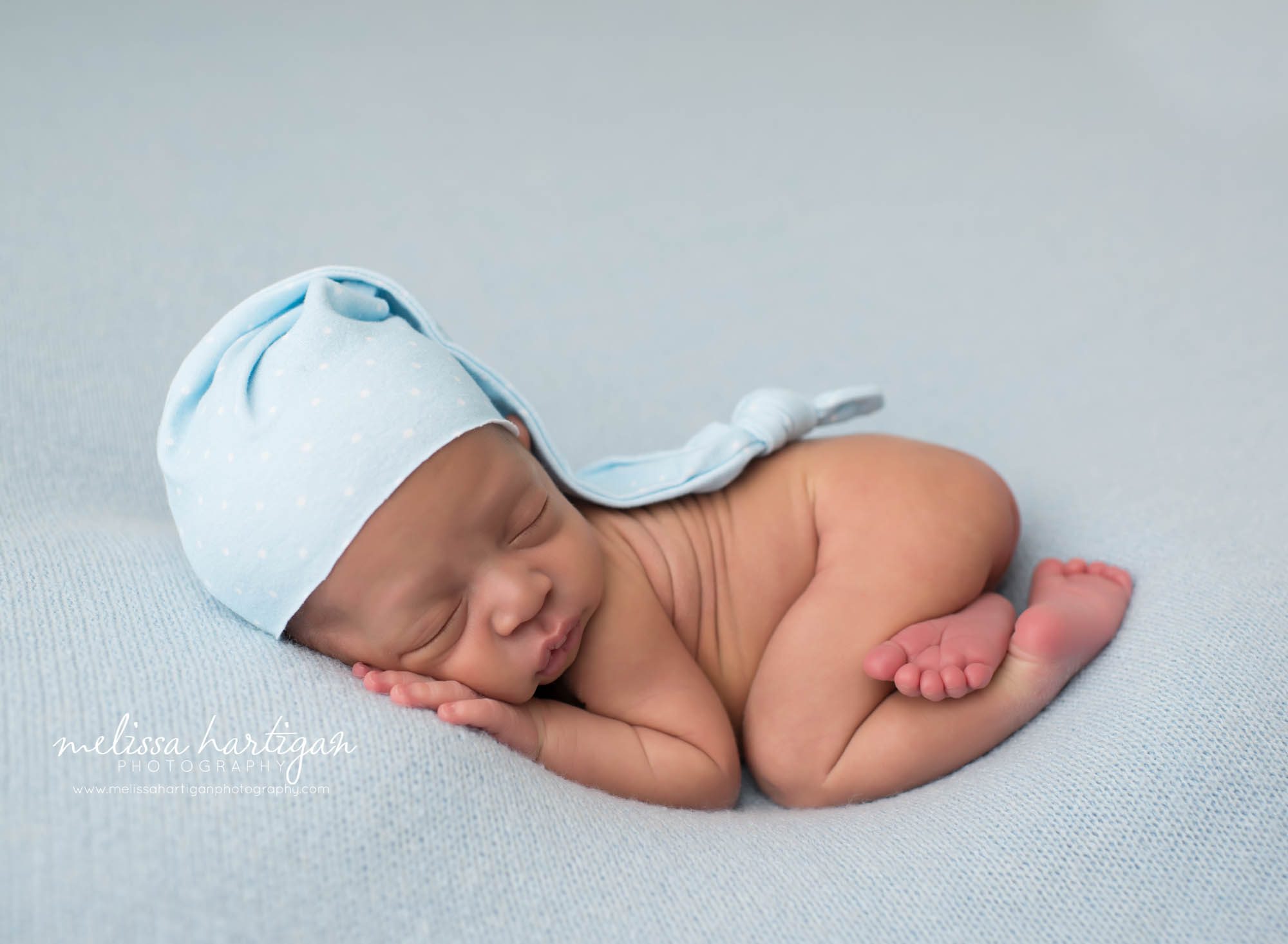newborn baby boy posed on blue backdrop with light blue sleepy cap with white polka dots