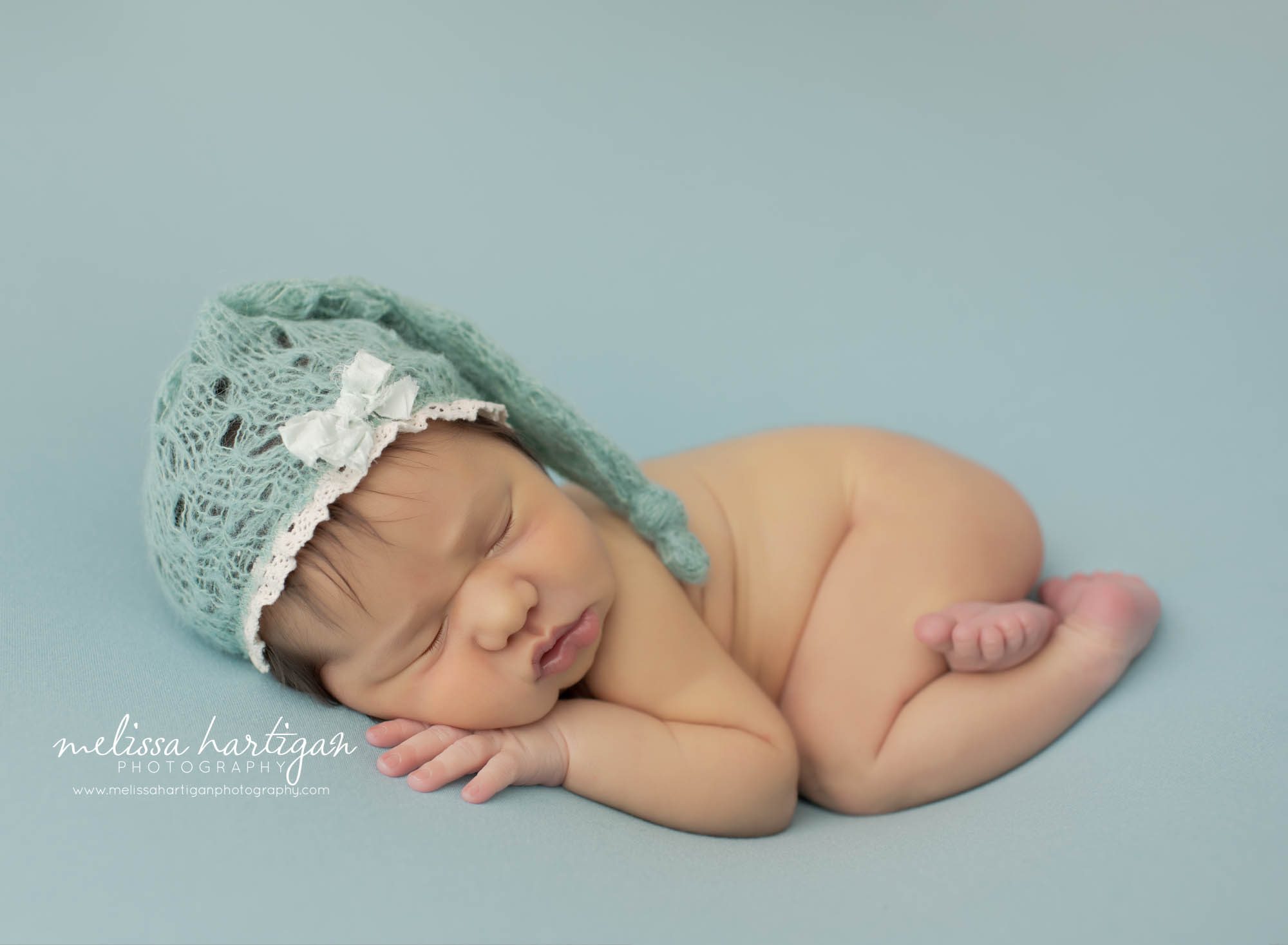 newborn baby girl posed on side wearing sea green knitted sleepy cap bonnet with bow