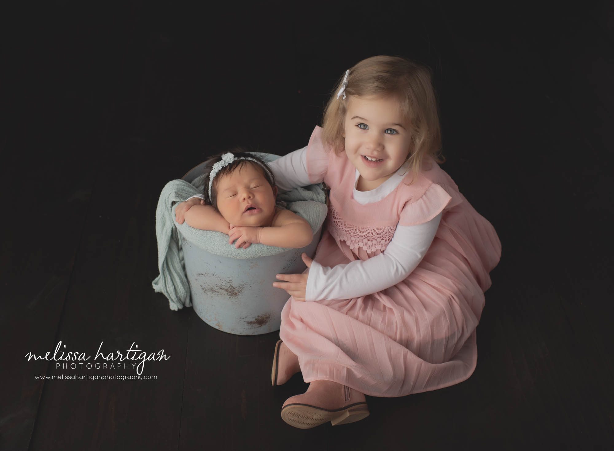 newborn baby girl posed in bucket with big sister holding her arm
