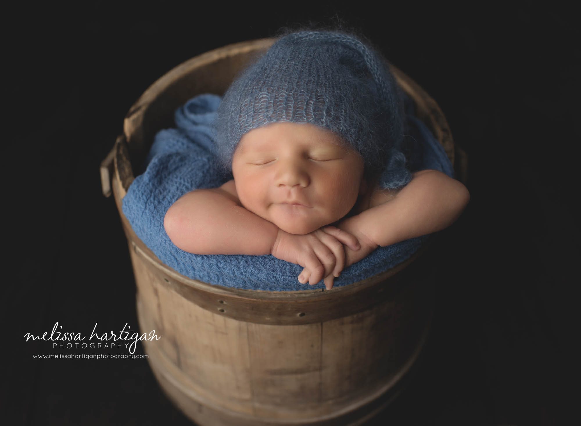 baby boy posed in wooden bucket with blue knitted sleepy cap smiling
