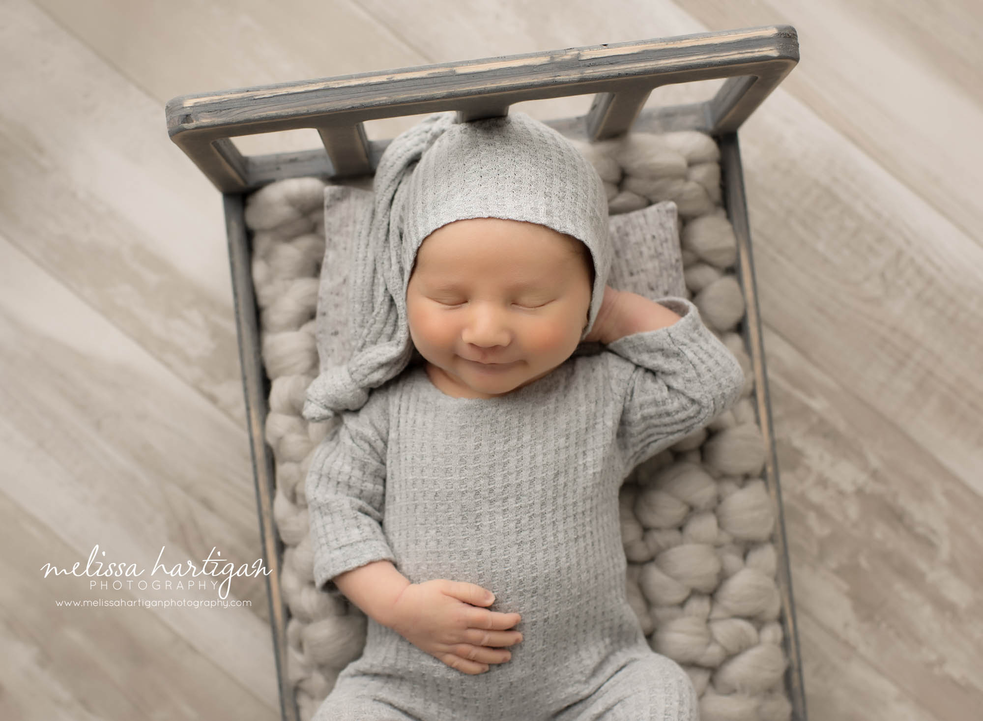 Newborn baby boy posed on back on wooden bed wearing grey waffled outfit and sleepy cap