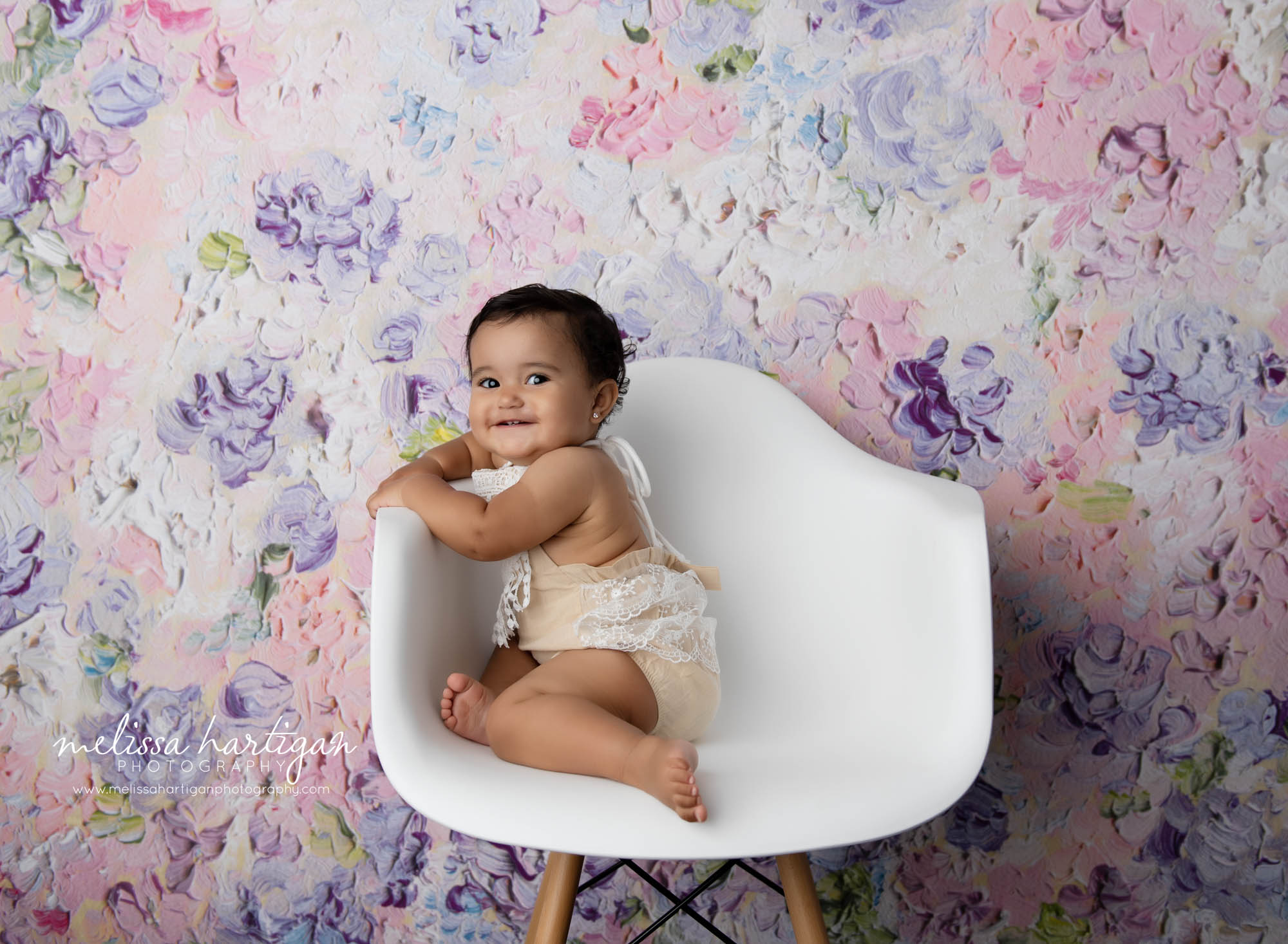 Baby girl sitting in white studio chair holding sides looking at camera smiling wearing baby milestone outfit