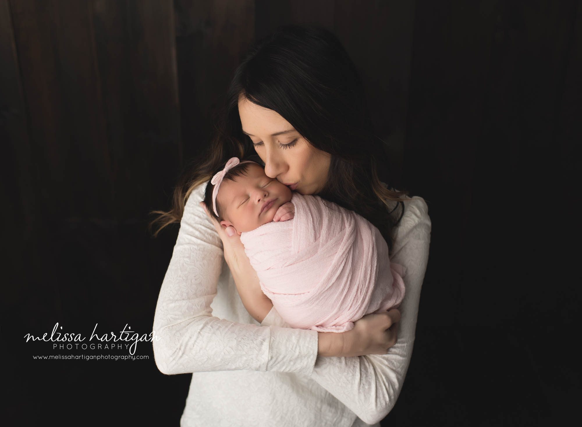mom kissing newborn baby girl wrapped in pink wrap with pink bow headband