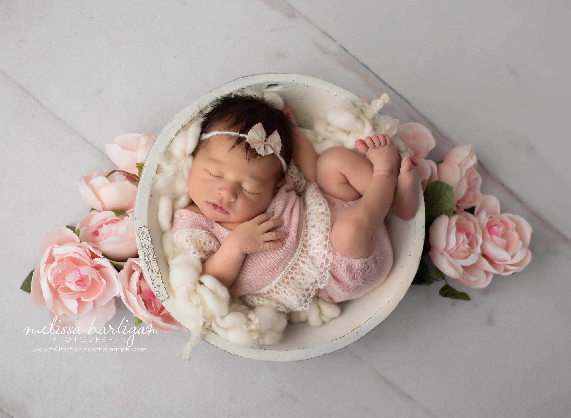 newborn baby girl posed in cream wooden bowl with pink flower bulbs pink knitted outfit and pink bow headband
