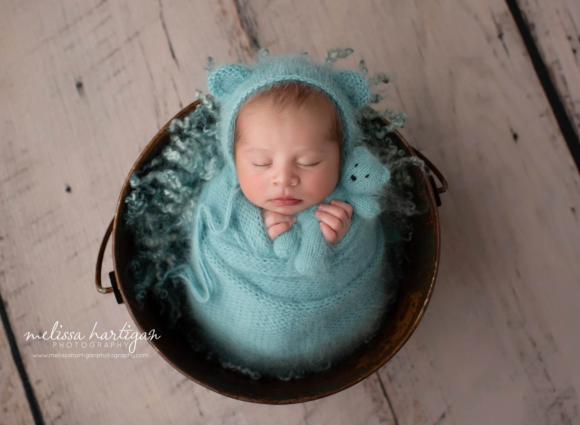 Newborn baby boy posed in bucket wearing teal colored knitted bear bonnet wrapped in knitted wrap with matching knitted teddy bear