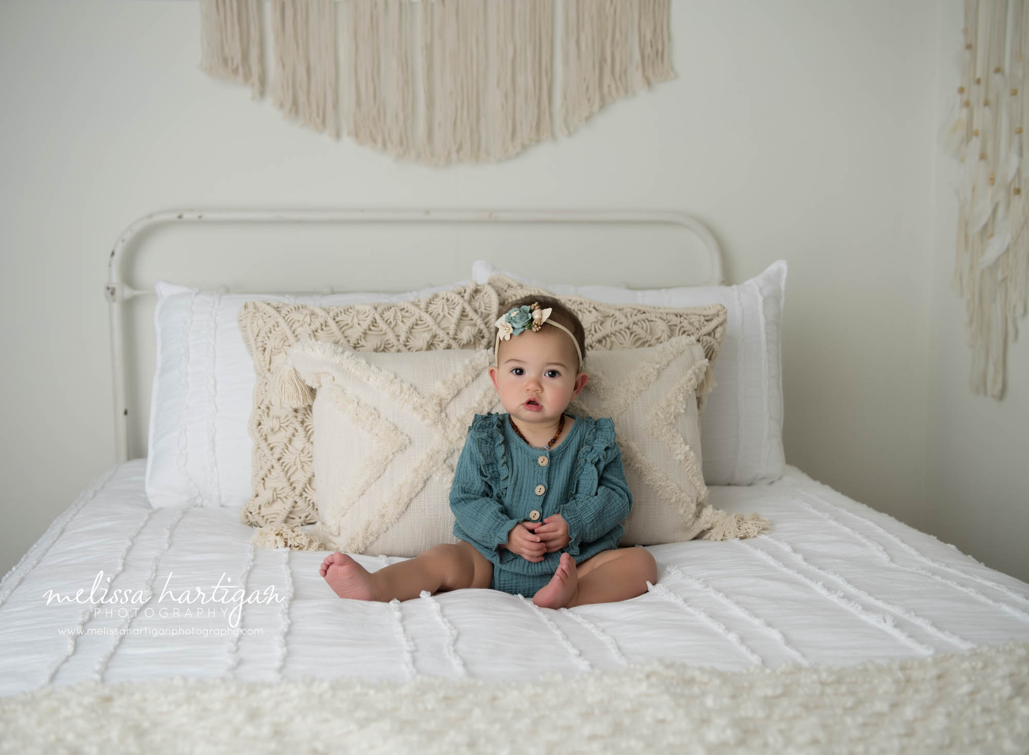 baby girl sitting on bed wearing teal baby milestone outfit with flower headband