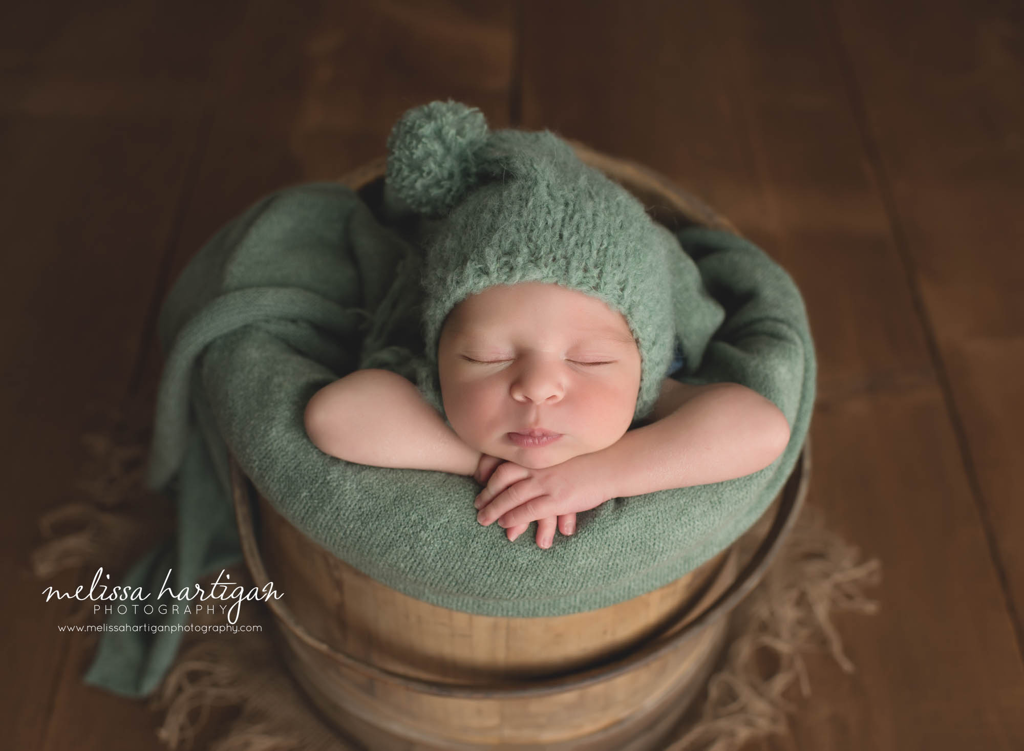 newborn baby boy wearing green knitted pom sleepy cap posed in wooden bucket