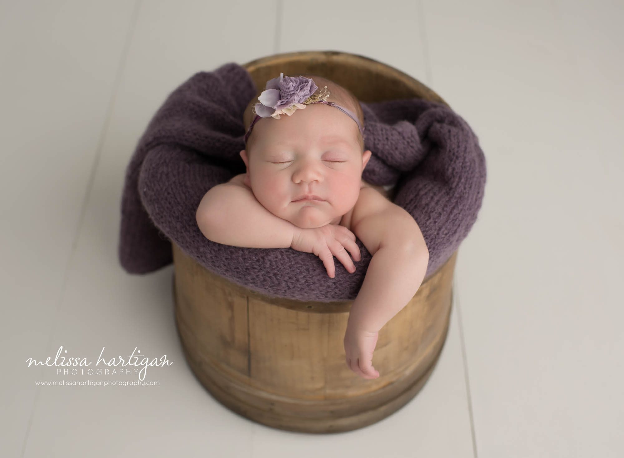newborn baby girl posed in wooden bucket with purple layer wrap and flower headband