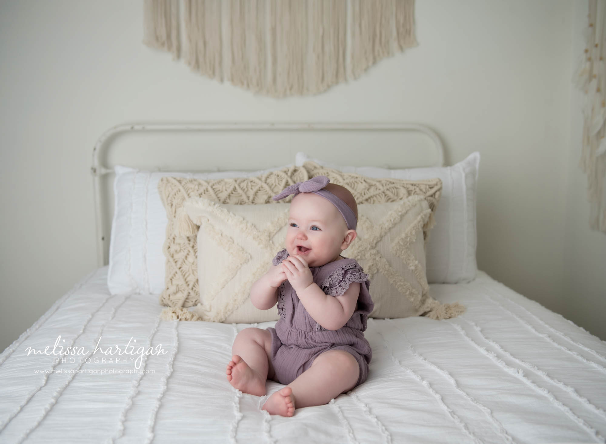 baby girl sitting on bed in CT baby photography studio with hands up to her face smiling and looking away