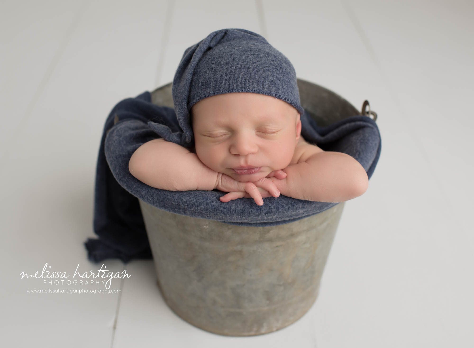 newborn baby boy posed in metal bucket with head on hands wearing navy blue sleepy cap CT newborn photography