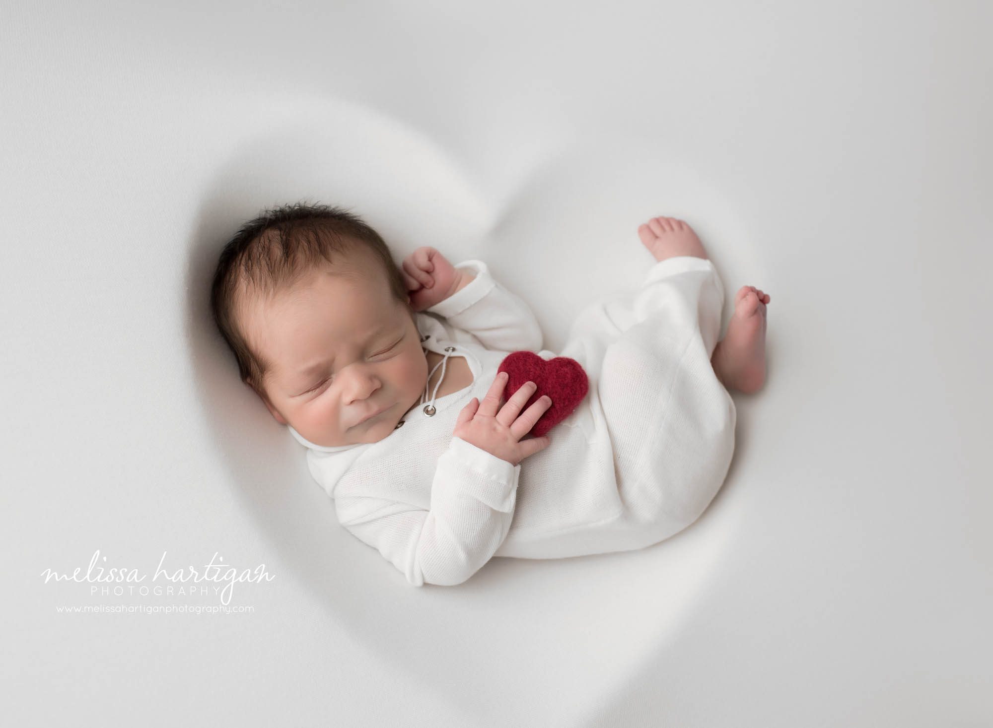 Newborn baby boy wearing white sleeper outfit posed on white backdrop with heart shaped outline underneath Hartford country Newborn Photography