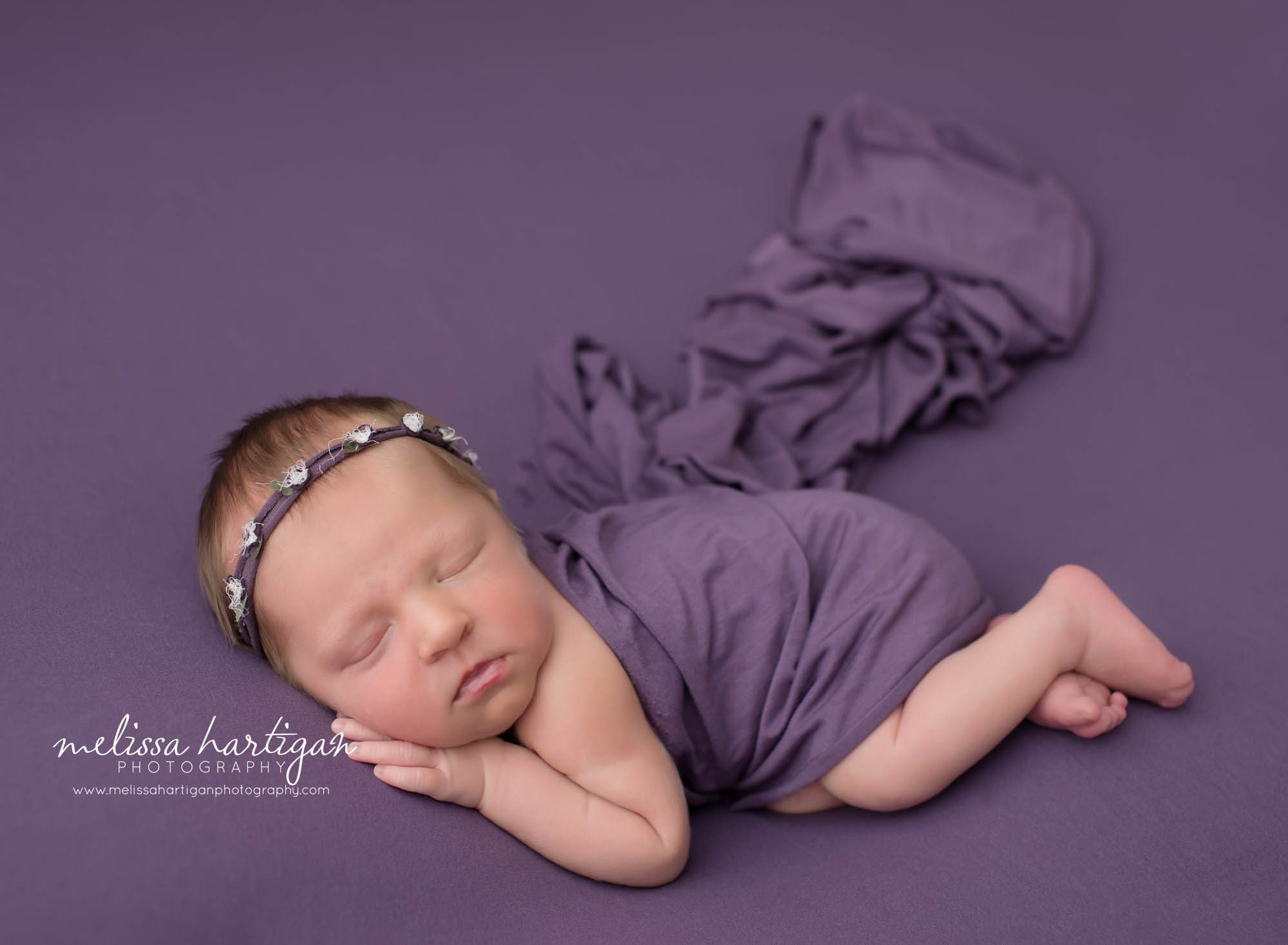 Baby girl posed on wide with hand under cheek purple backdrop with matching purple wrap and purple flower headband