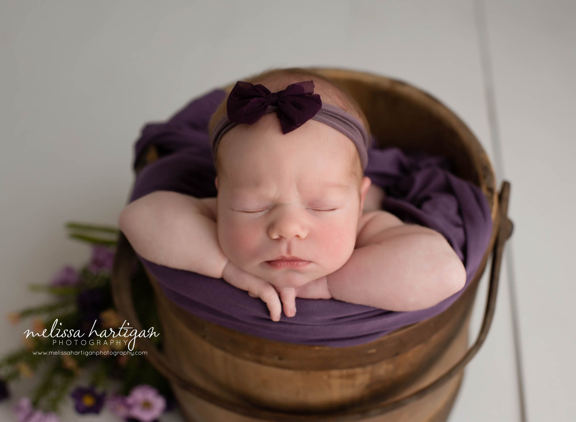 Newborn baby girl posed in wooden bucket with purple flower elements and purple bow headband
