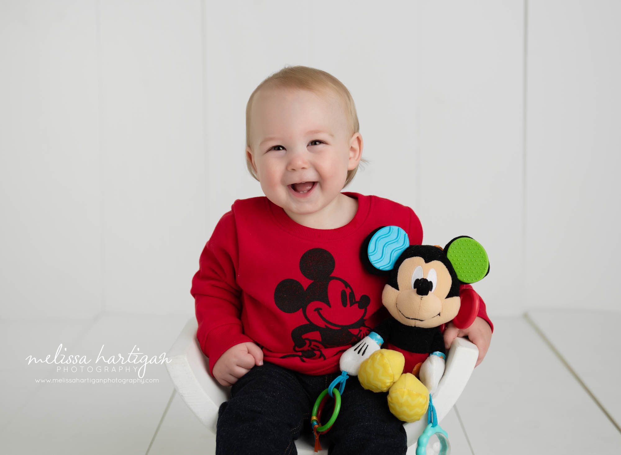 baby boy holding mickey mouse stuffy sitting on white wooden chair in photography studio melissa hartigan photography