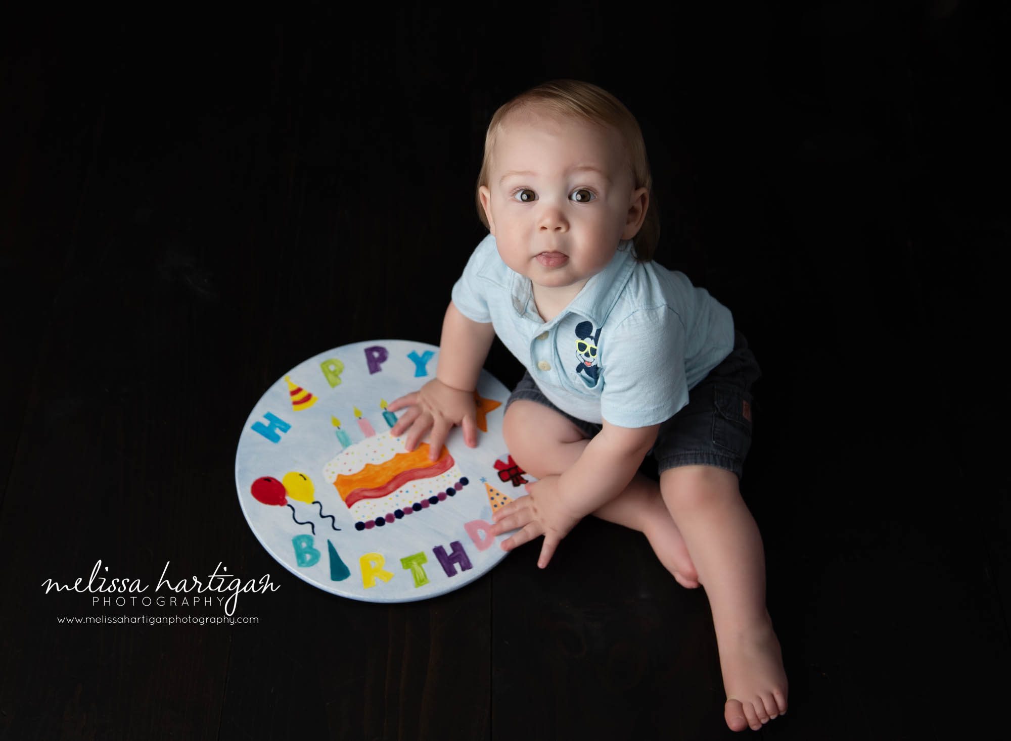 baby boy sitting on wooden backdrop with happy birthday sign in hands looking up at camera baby photography columbia ct