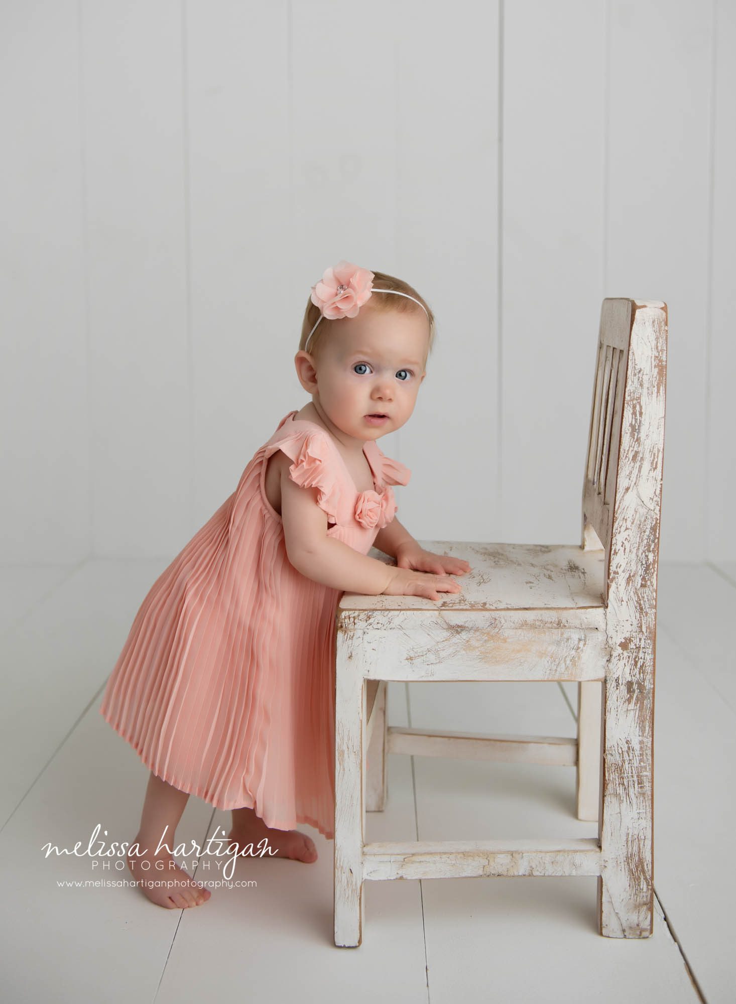 baby girl standing by wooden white chair wearing coral pink dress and matching headband Colchester CT baby Photography