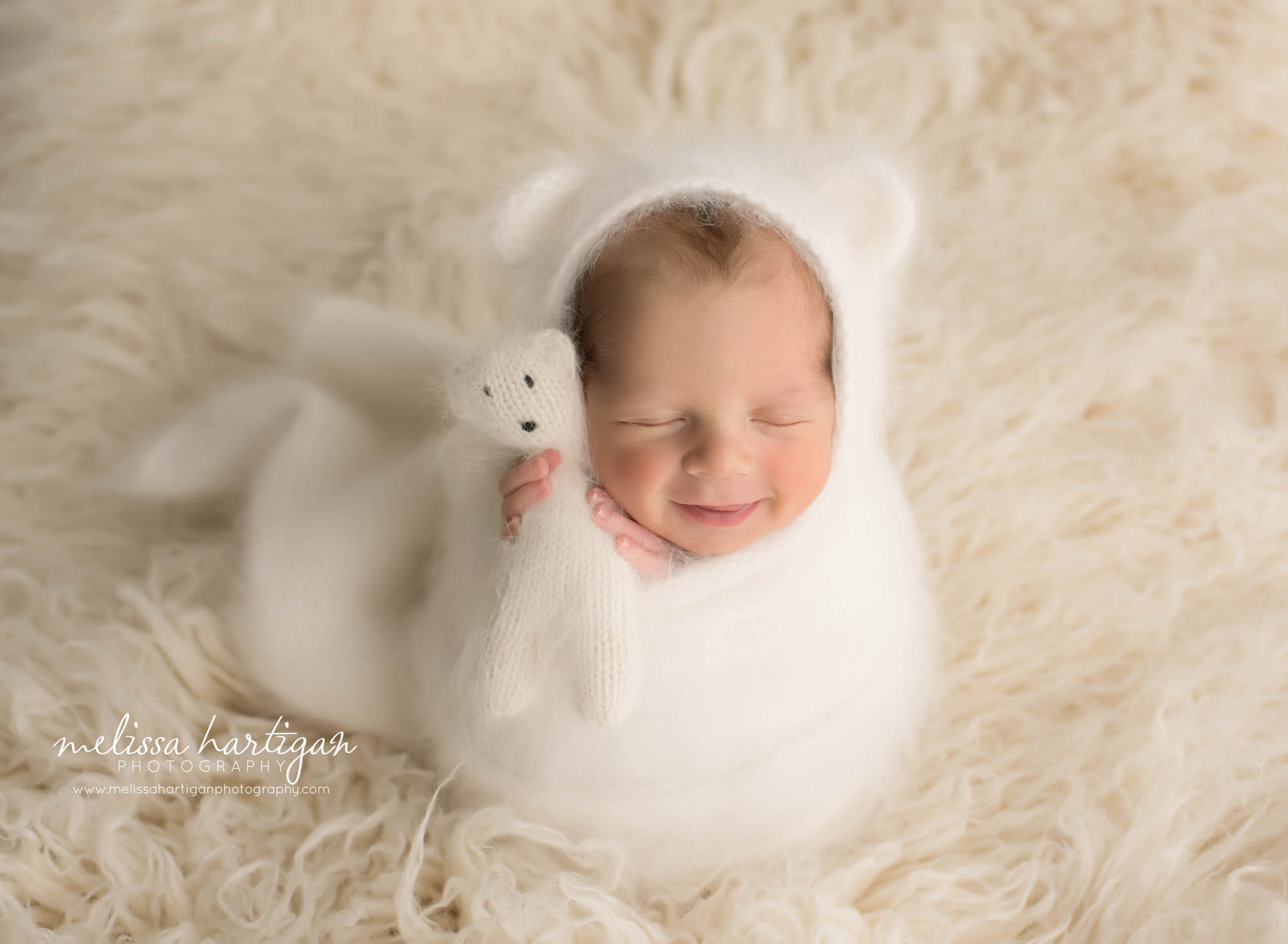 Newborn baby boy smiling in posed newborn photography picture wrapped in white knitted wrap with matching angora bear bonnet and matching teddy