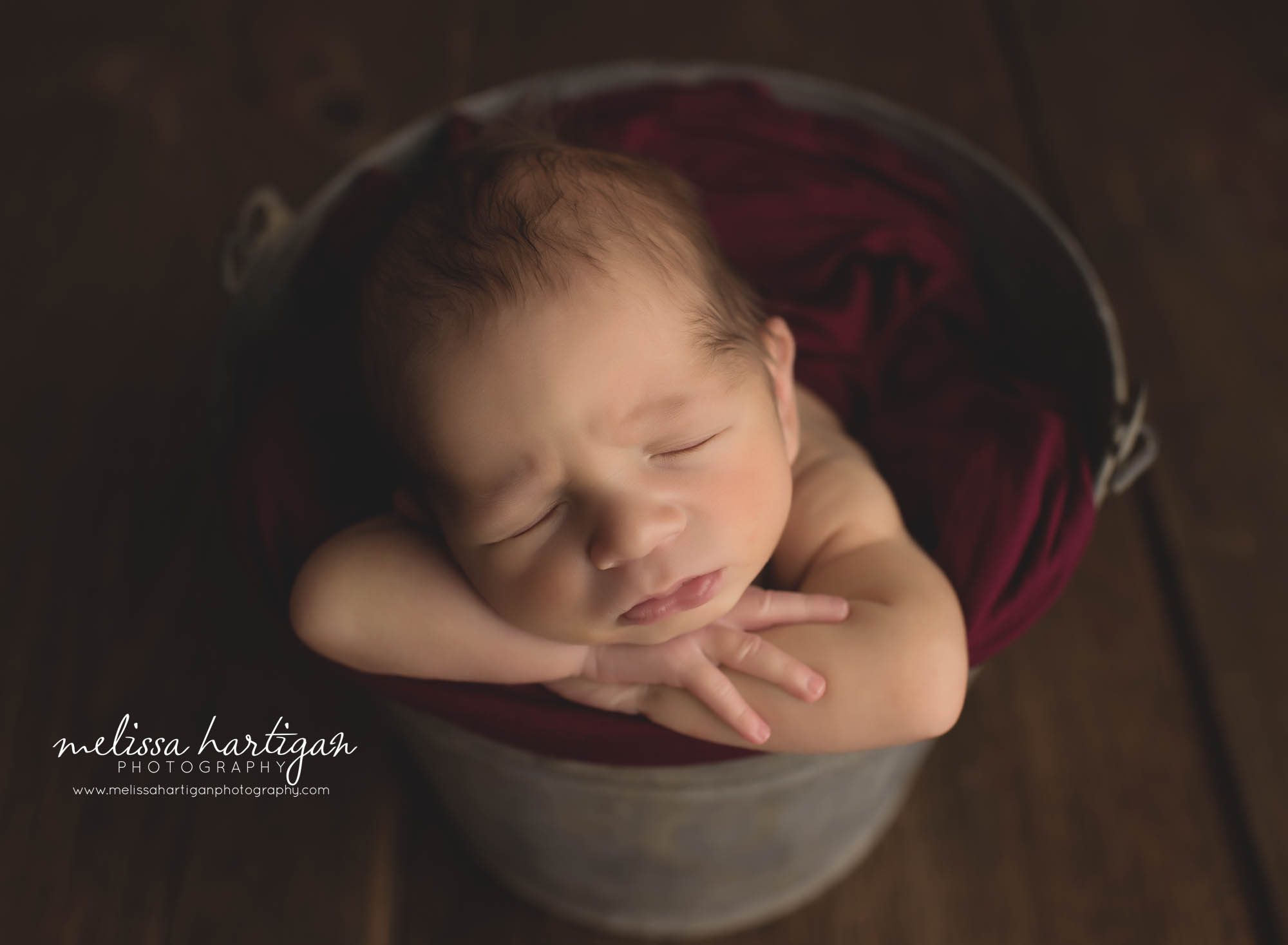 newborn posed with chin on hands in metal bucket with burgundy color wrap