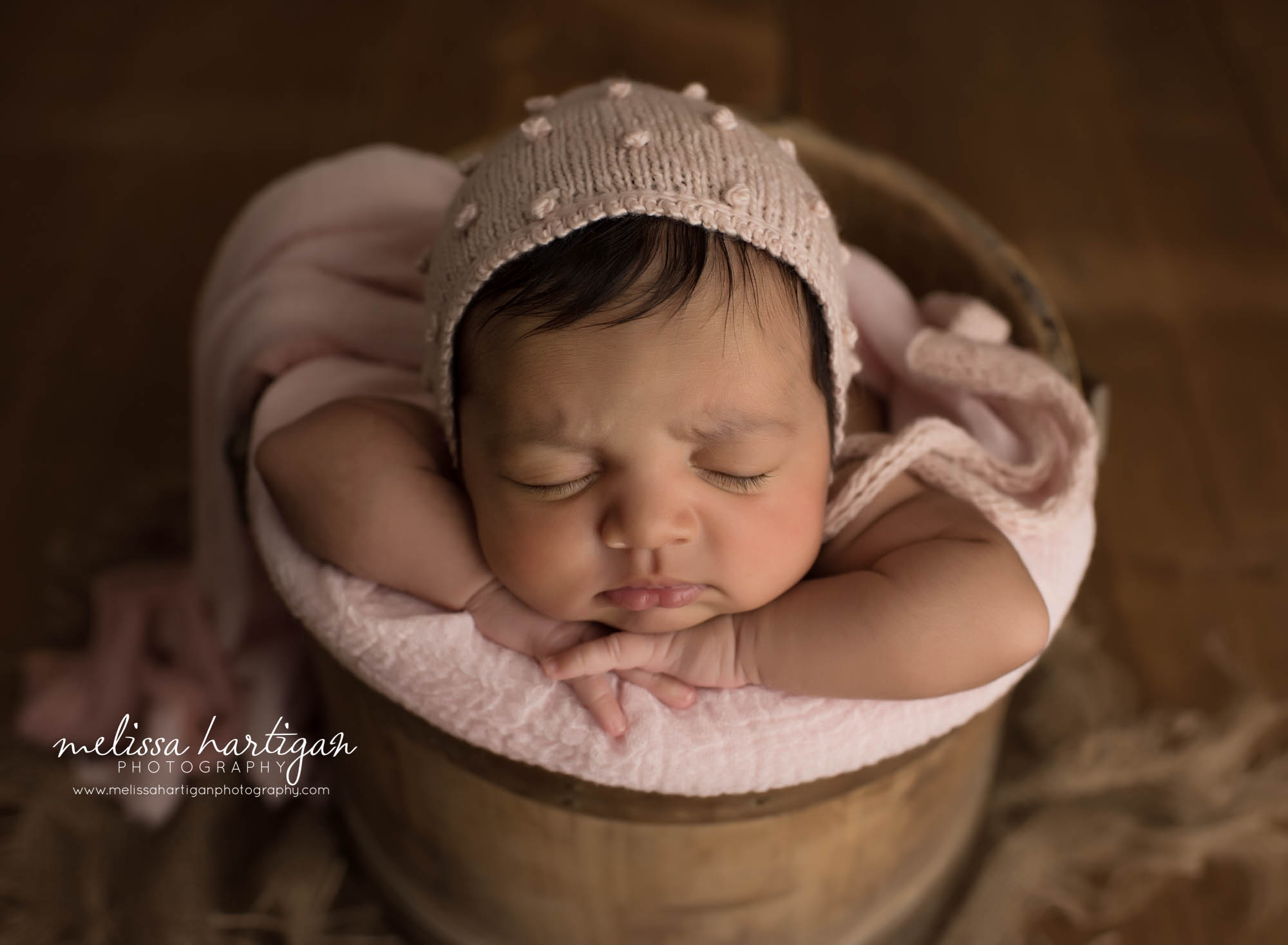 newborn baby girl posed with chin on hands in wooden barrel bucket with light pink knitted bonnet