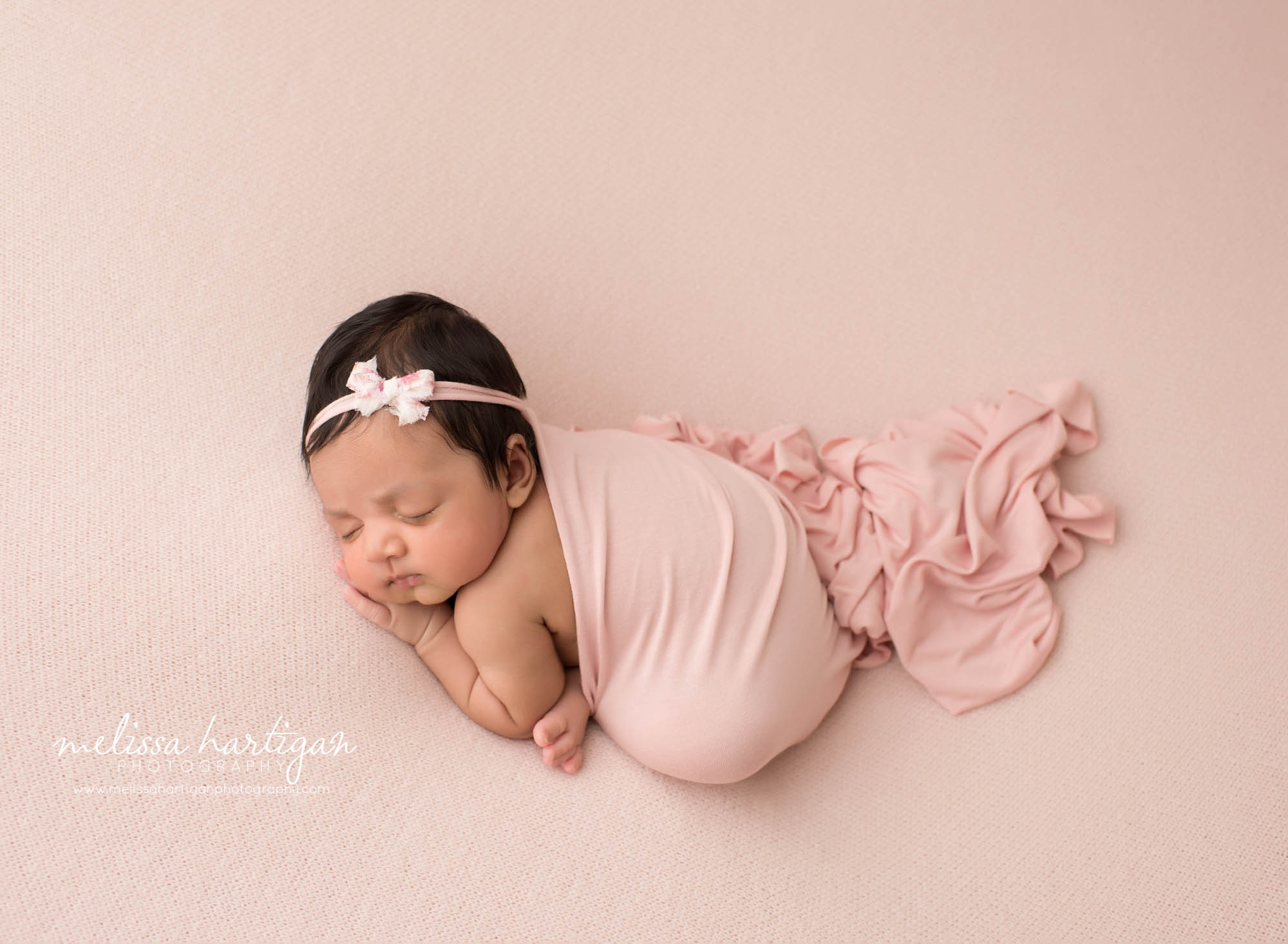 newborn baby girl posed on pink backdrop with bow headband and [ink wrap draped over