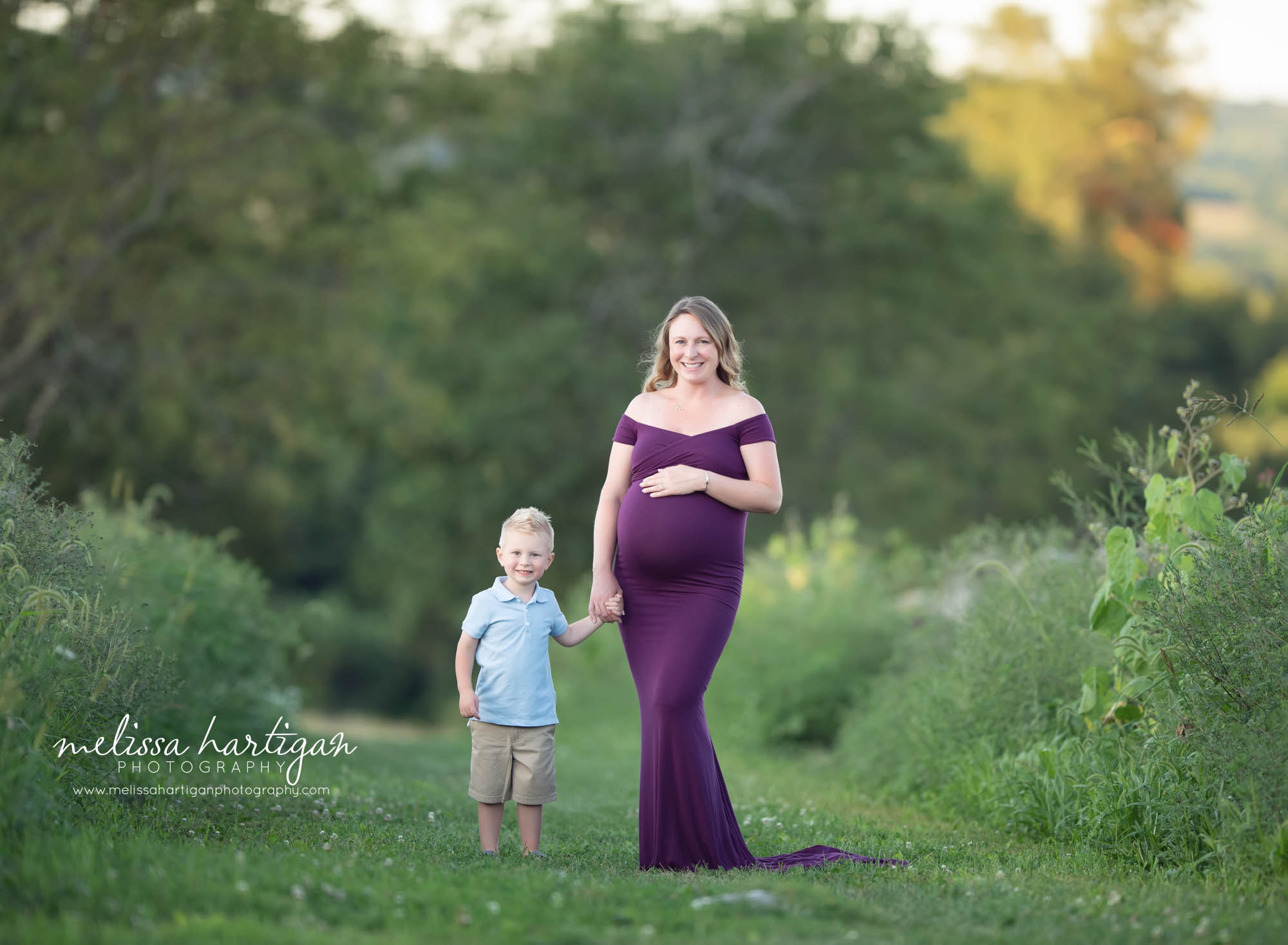 mom holding toddler son's hand and holding baby bump wearing long form fitting purple maternity dress