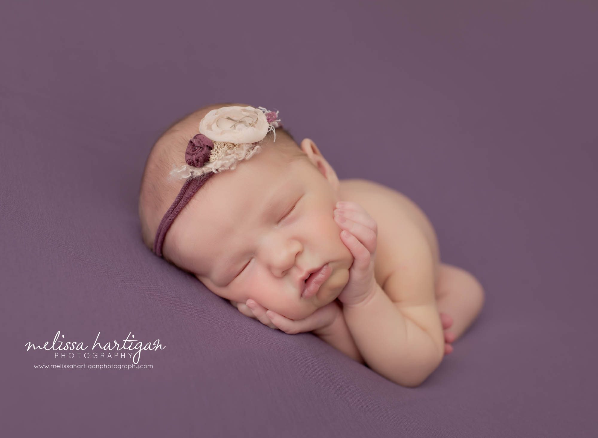 newborn baby girl posed on side on purple backdrop with pretty headband