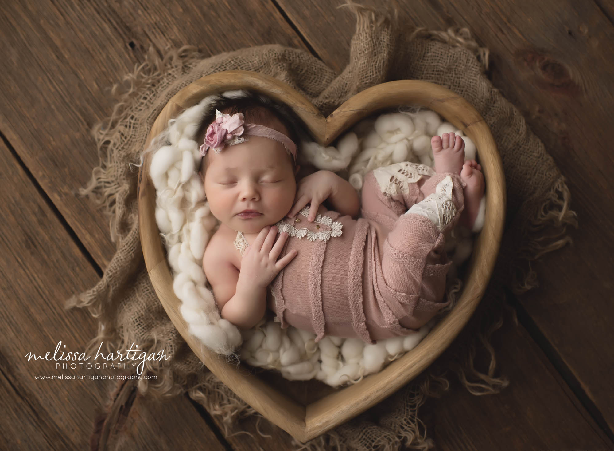 newborn baby girl posed in heart shaped wooden prop with pink outfit and flower headband