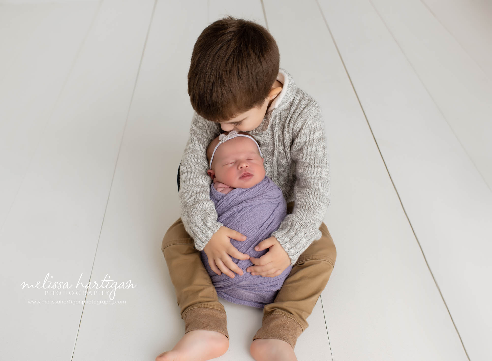 toddler big brother sitting on floor with newborn baby sister between his legs giving her a kiss on her head