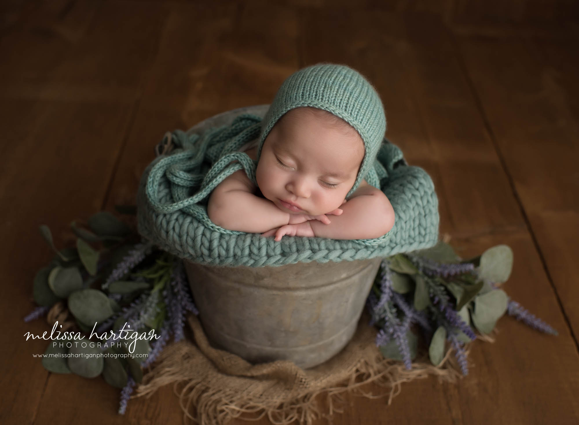 newborn baby girl posed in metal bucket with light colored teal knitted bonnet and wrap