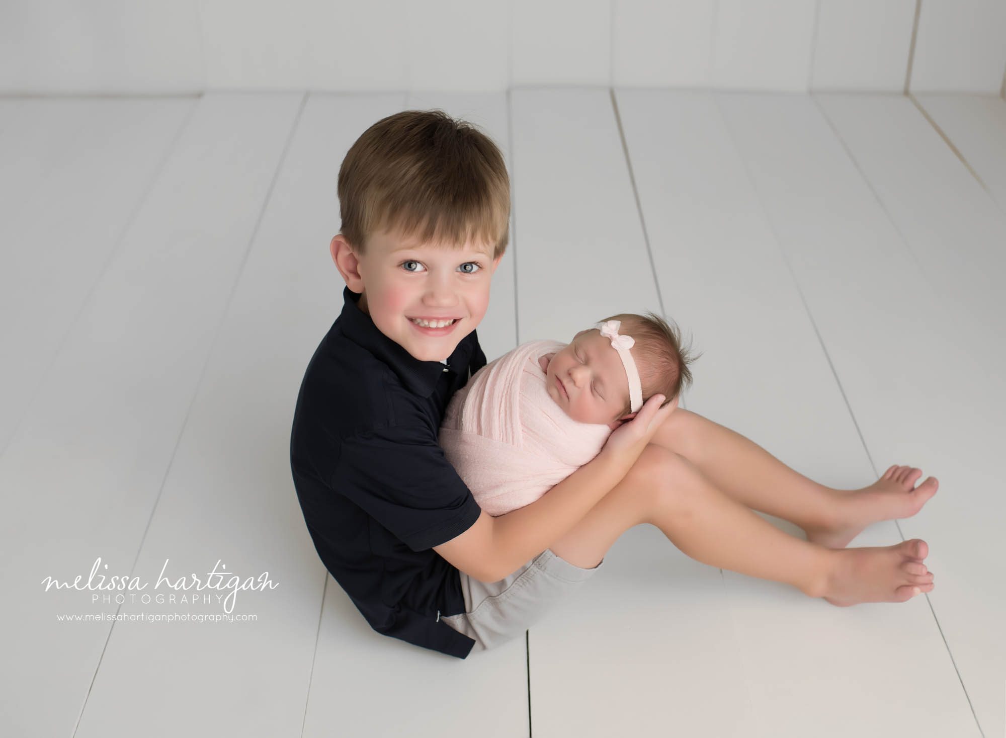 big brother sitting on wooden planks in connecticut photography studio holding newborn baby sister for family photo