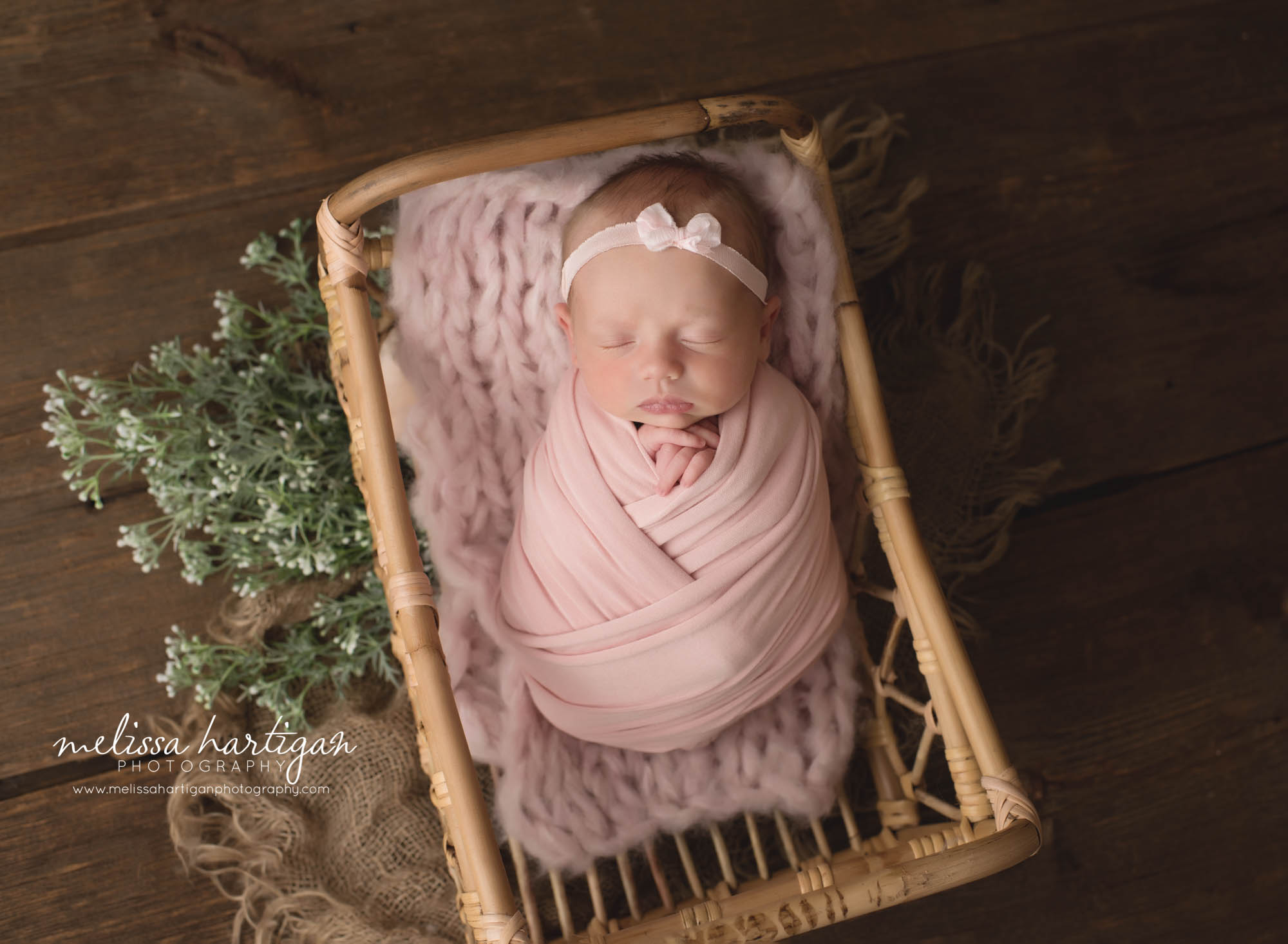 newborn baby girl wrapped in light pink wrap with matching pink bow headband posed in basket with green foilage hartford county newborn baby photographer