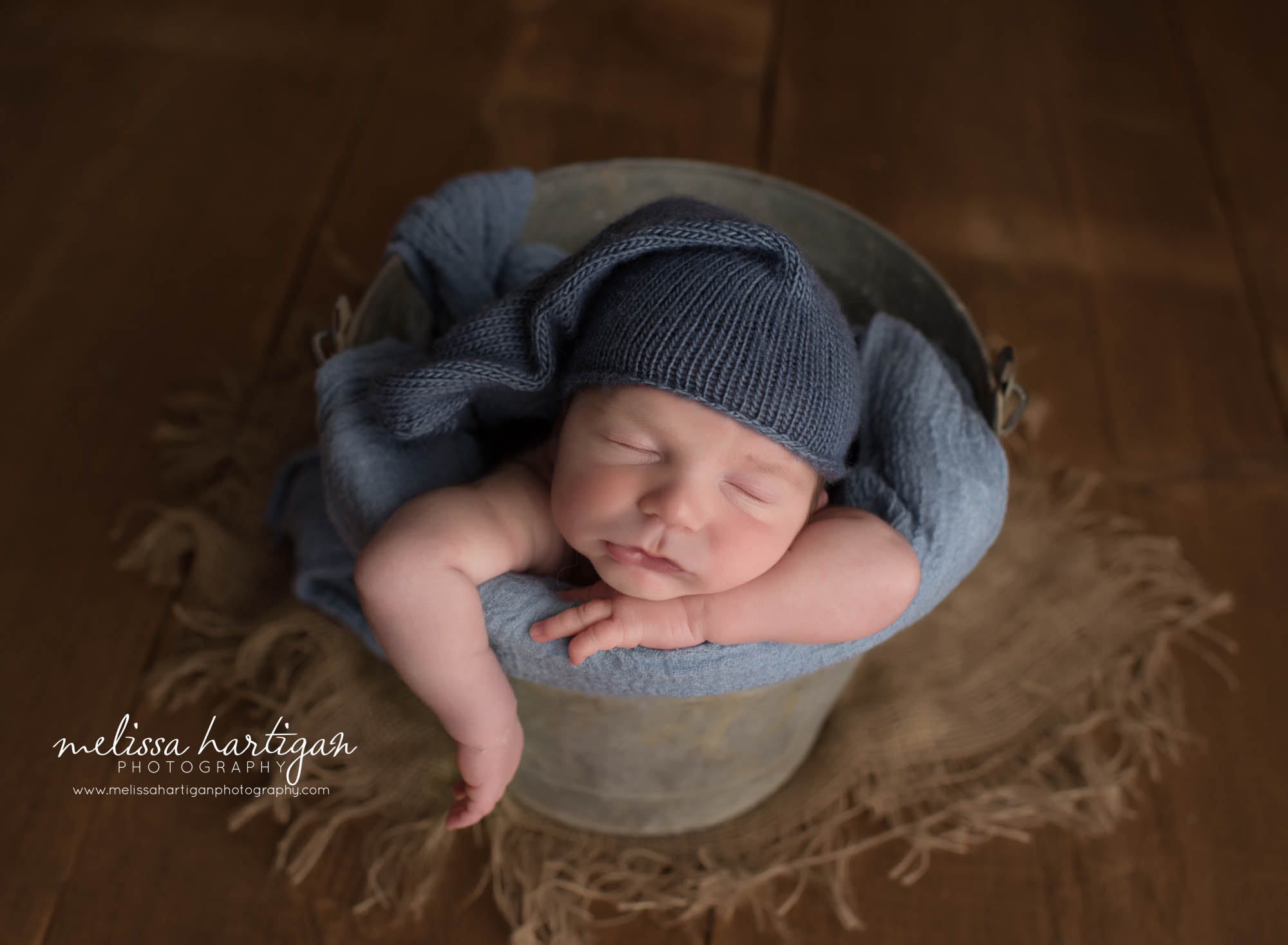 newborn boy posed in bucket with arm handing out sleeping blue knitted sleepy cap on