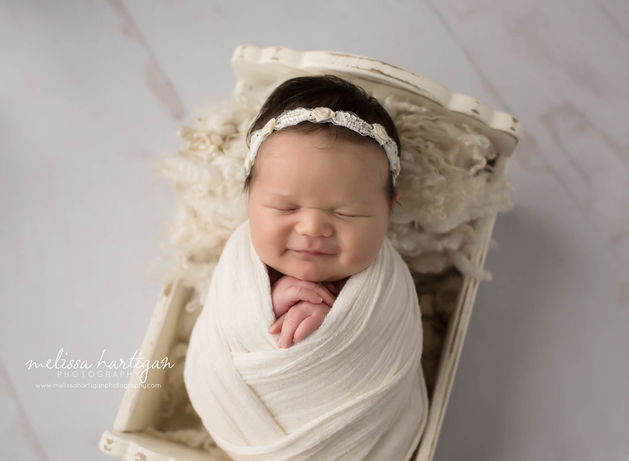 Newborn baby girl smiling wrappe din cream wrap posed in wooden newborn craddle prop