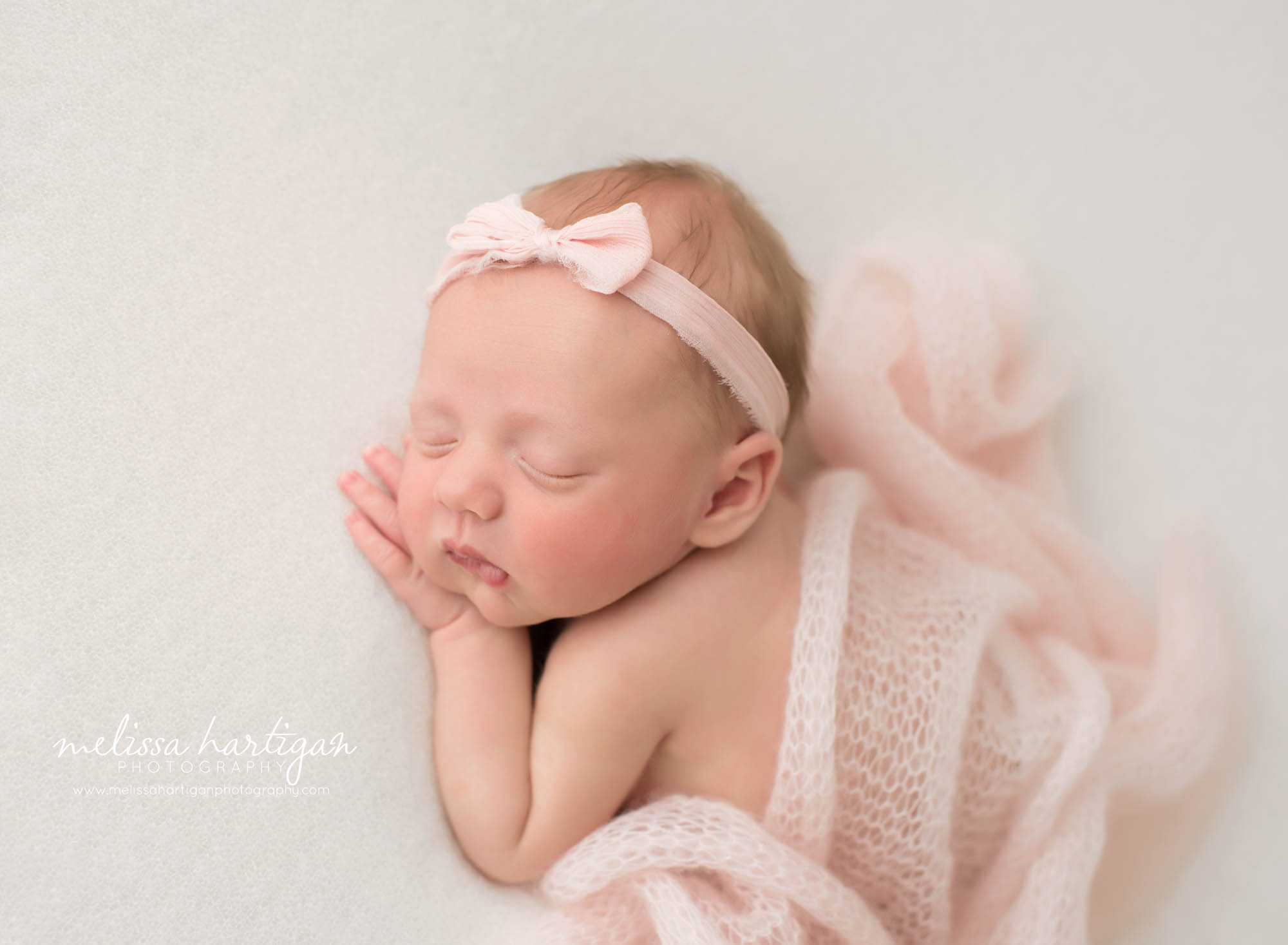 newborn baby girl pose don tummy with hand under cheek with light pink knitted layer drapped over her back wearing light pink bow headband newborn photography hartford county ct