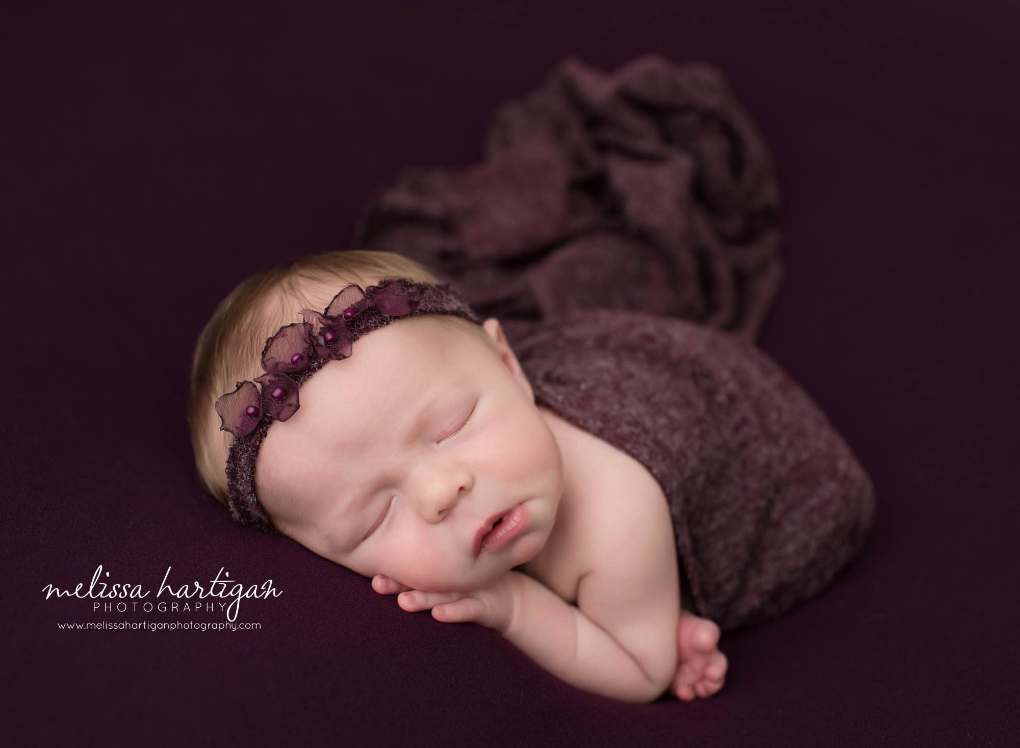 newborn baby girl posed on eggplant purple backdrop with headband and wrap draped over